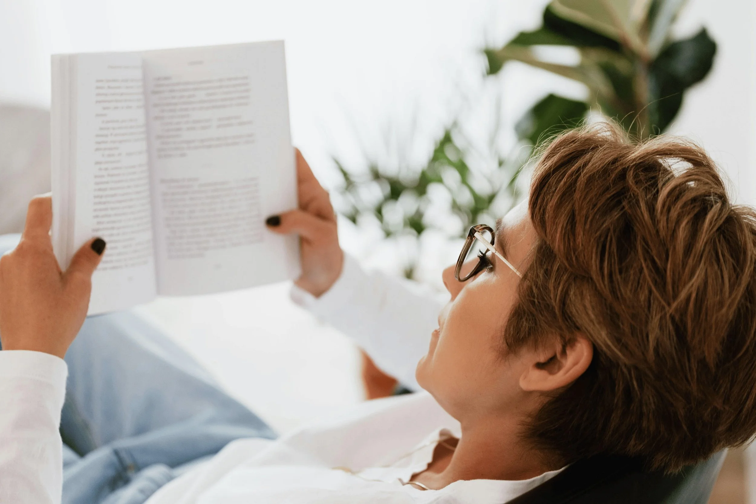 A woman with short brown hair and glasses is lying on her the couch reading a book, enjoying the time returned by Lighter Home household management service in Gainesville FL. There is a large green houseplant in the background.