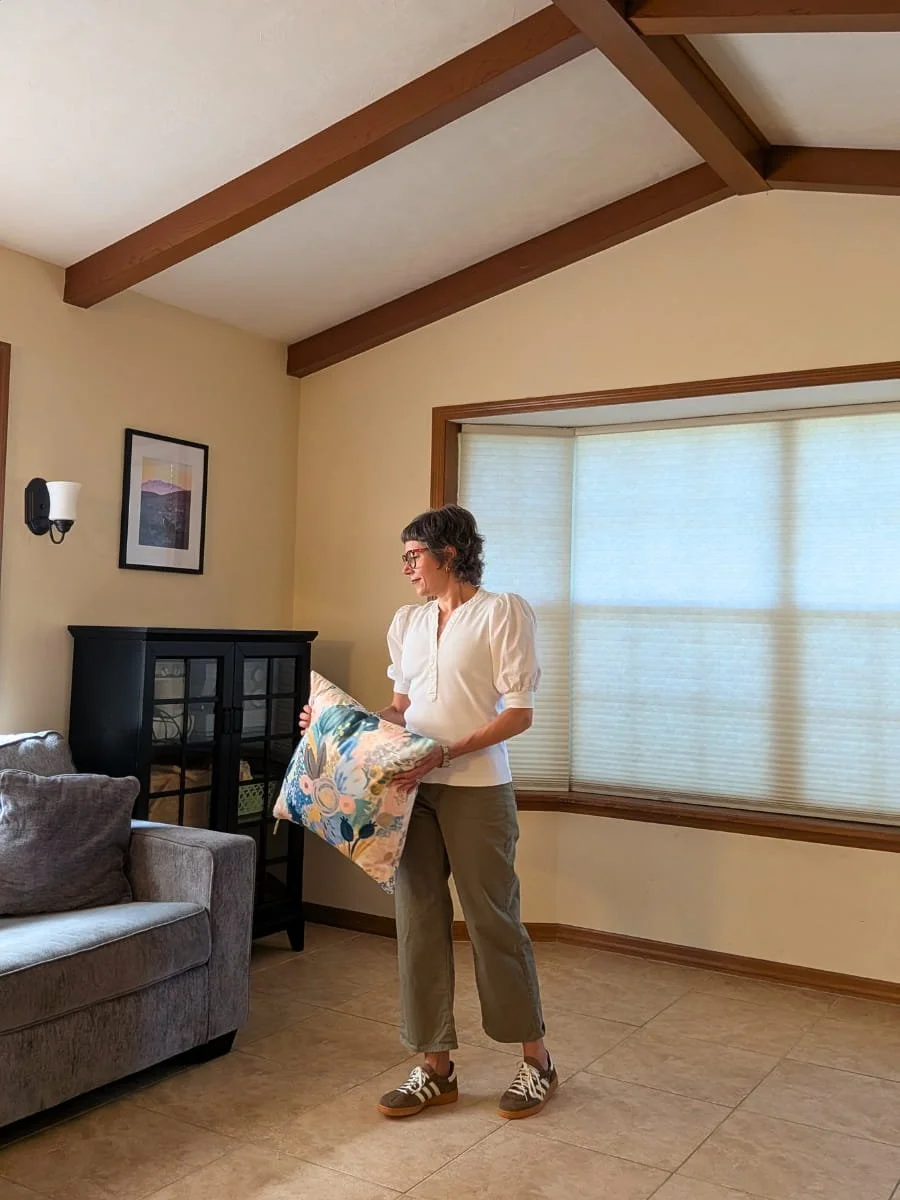 Colleen Raccioppi, founder of Lighter Home, resetting a living room room that features beige walls, wooden beams on the ceiling, a gray sofa with pillows, a black cabinet with glass doors, and two windows with beige blinds.