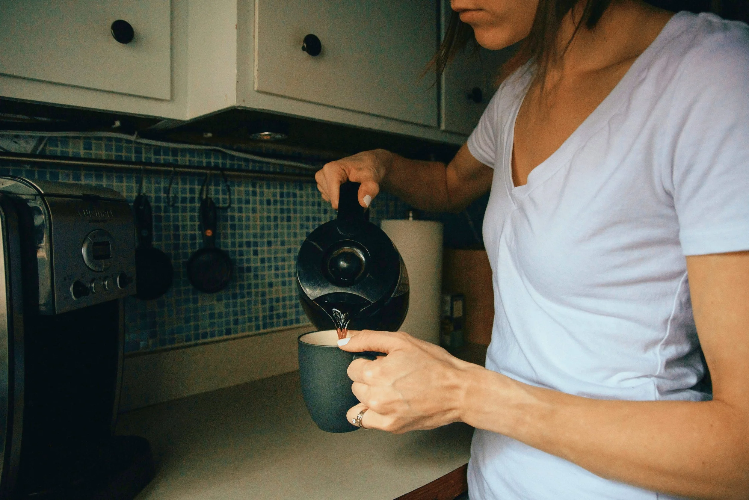 A tired woman in a white T-shirt pouring coffee from a French press into a mug in a kitchen with white cabinets and a blue tiled backsplash. Lighter Home household management service reduces cognitive labor for busy parents.