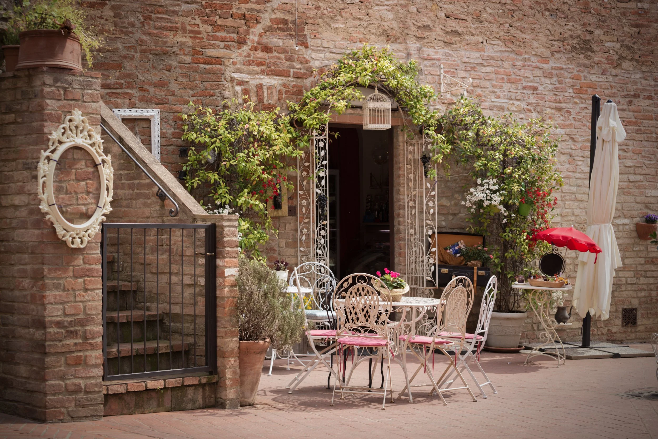 A courtyard with a brick wall, green plants, and outdoor furniture including white wrought iron chairs and tables, a red umbrella, and decorative items.