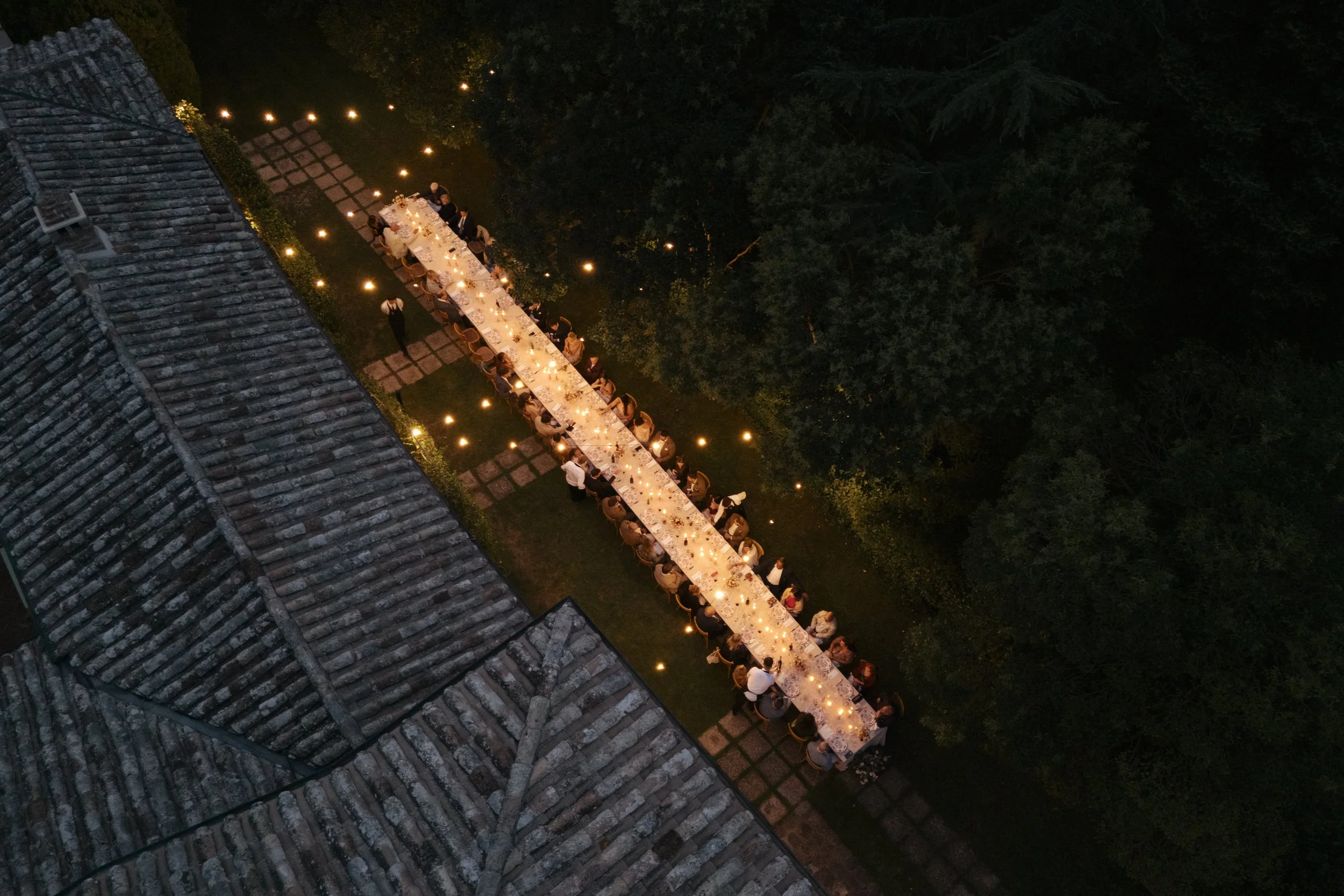 A long outdoor dinner table set with candles and surrounded by people, illuminated by string lights, in a lush garden area at night.