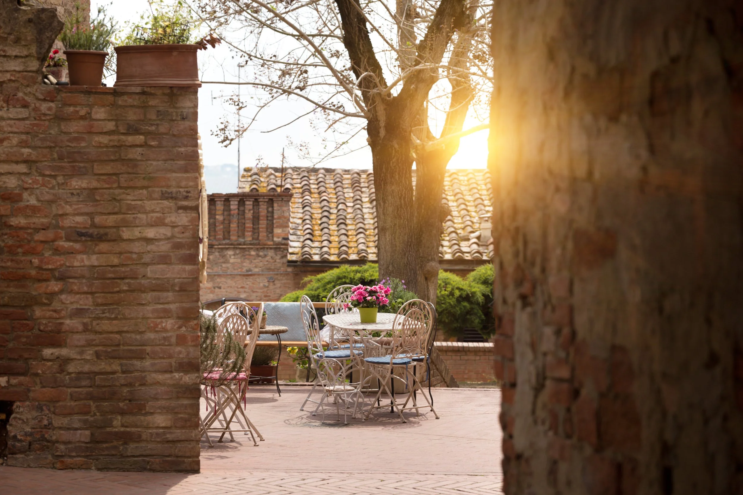 A charming outdoor patio with white wrought iron chairs and a table, decorated with a pot of pink flowers, surrounded by brick walls and a large tree, with sunlight filtering through the branches.