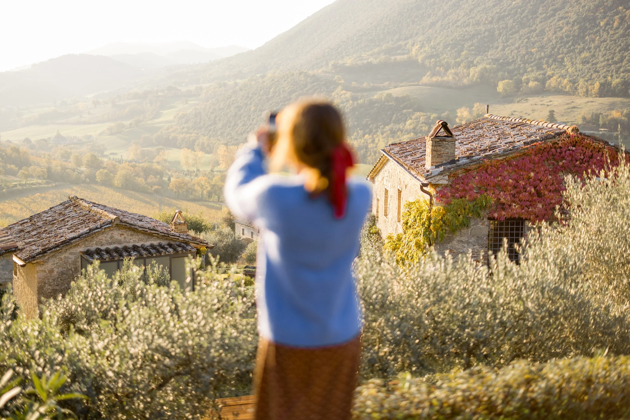 A person taking a photo with a smartphone on a balcony overlooking a rural landscape with houses, trees, and mountains in the distance during daylight.
