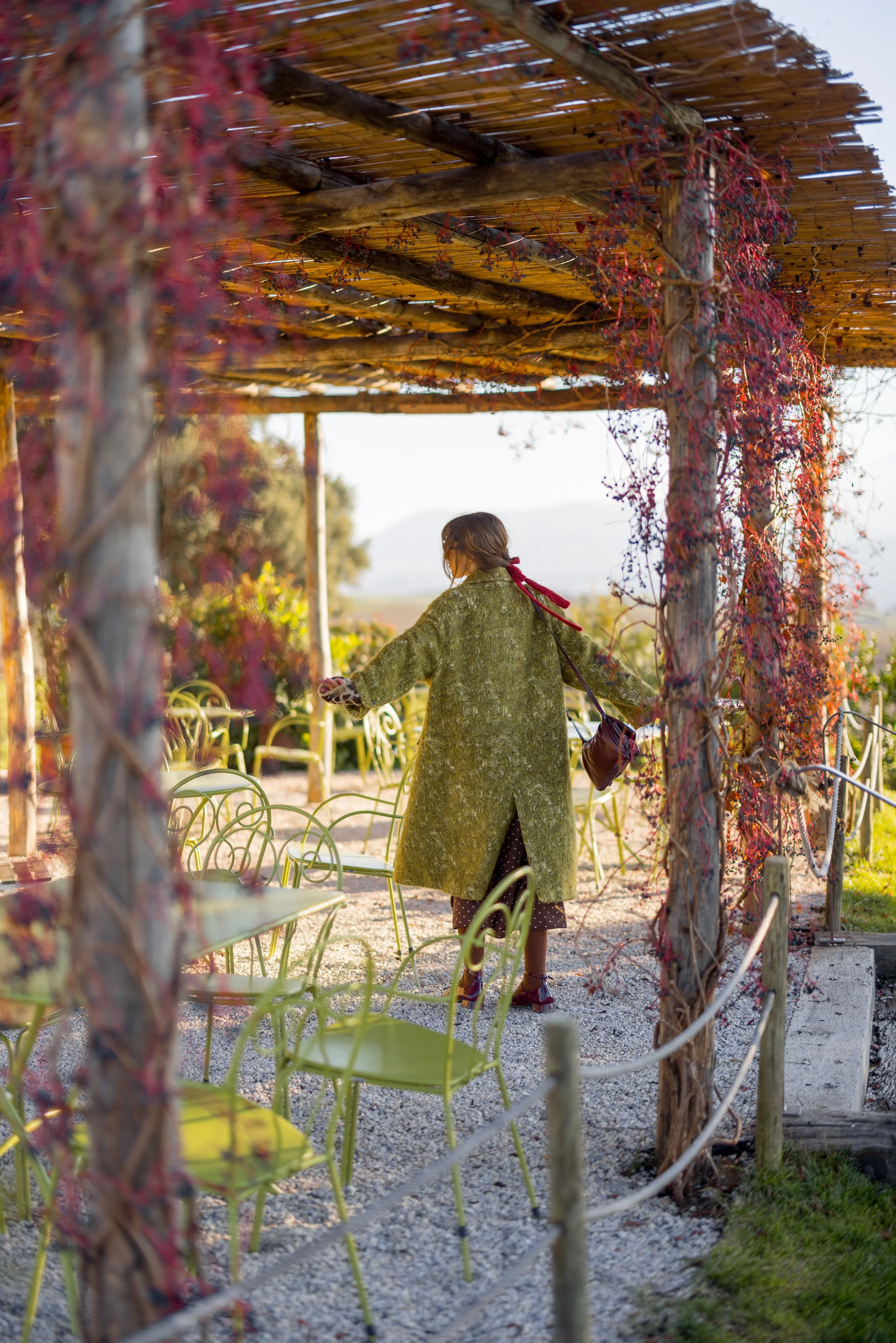 A woman in a green coat and polka dot dress standing in a garden under a wooden pergola covered with red, dried vines. She holds a phone in one hand and a bag in the other, surrounded by yellow chairs and lush greenery.