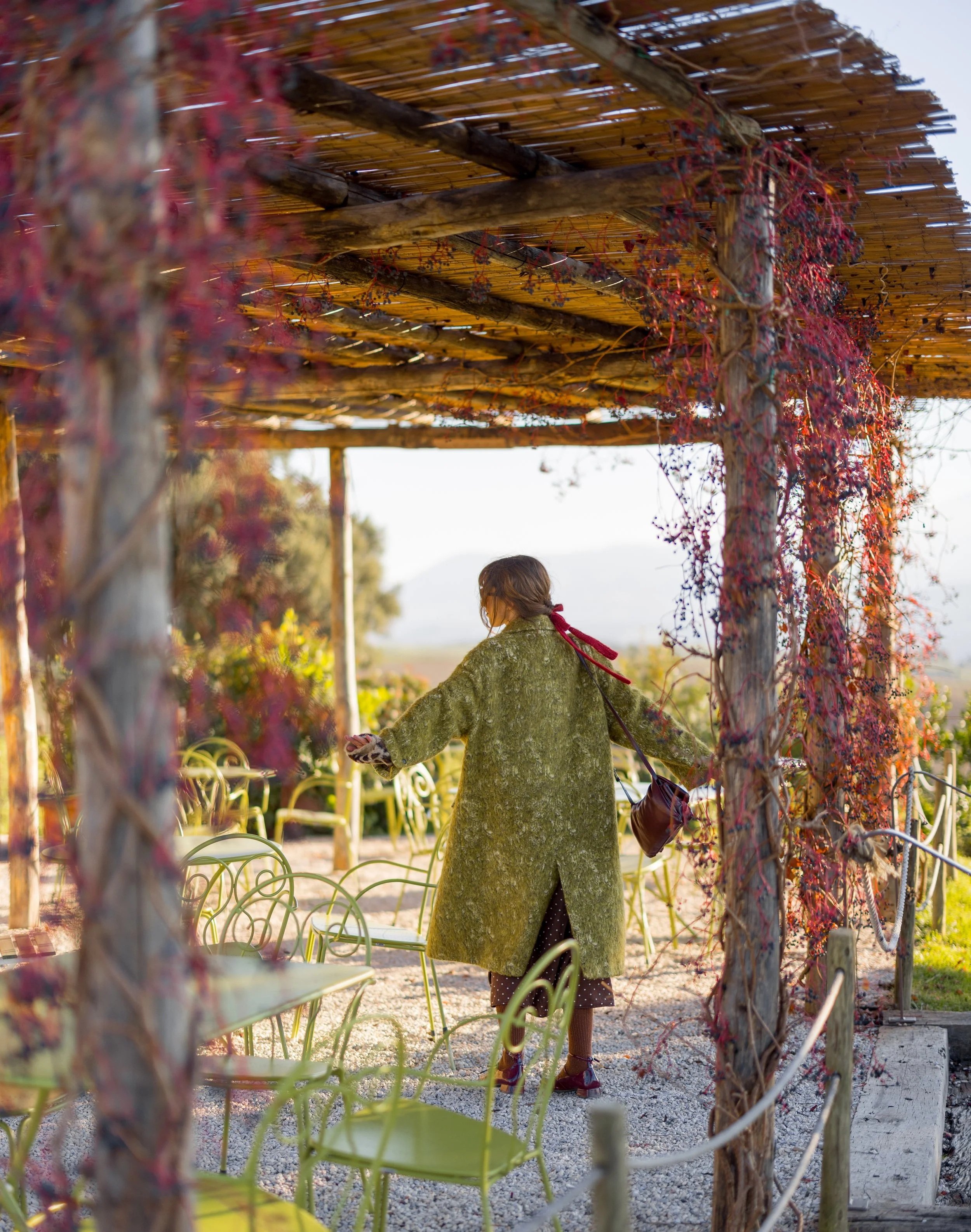 A woman in a green coat and polka dot skirt stands under a rustic wooden pergola with vine-covered beams, in an outdoor garden setting with chairs, plants, and trees in the background.