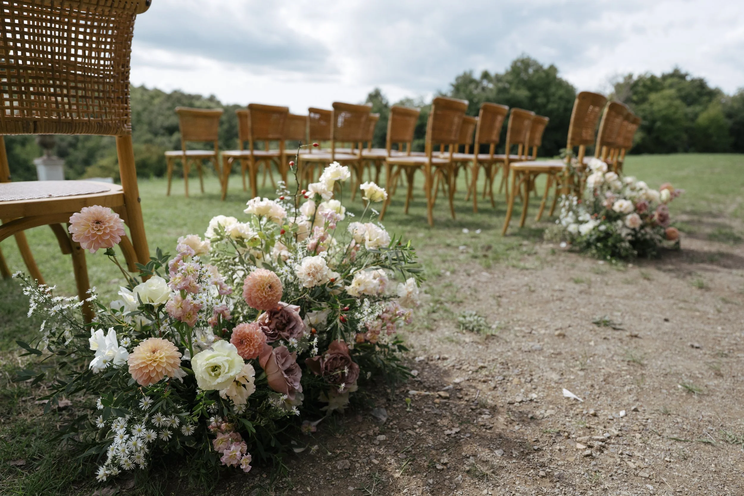 Outdoor wedding setup with a row of wooden chairs and floral arrangements on a grassy field under a cloudy sky.