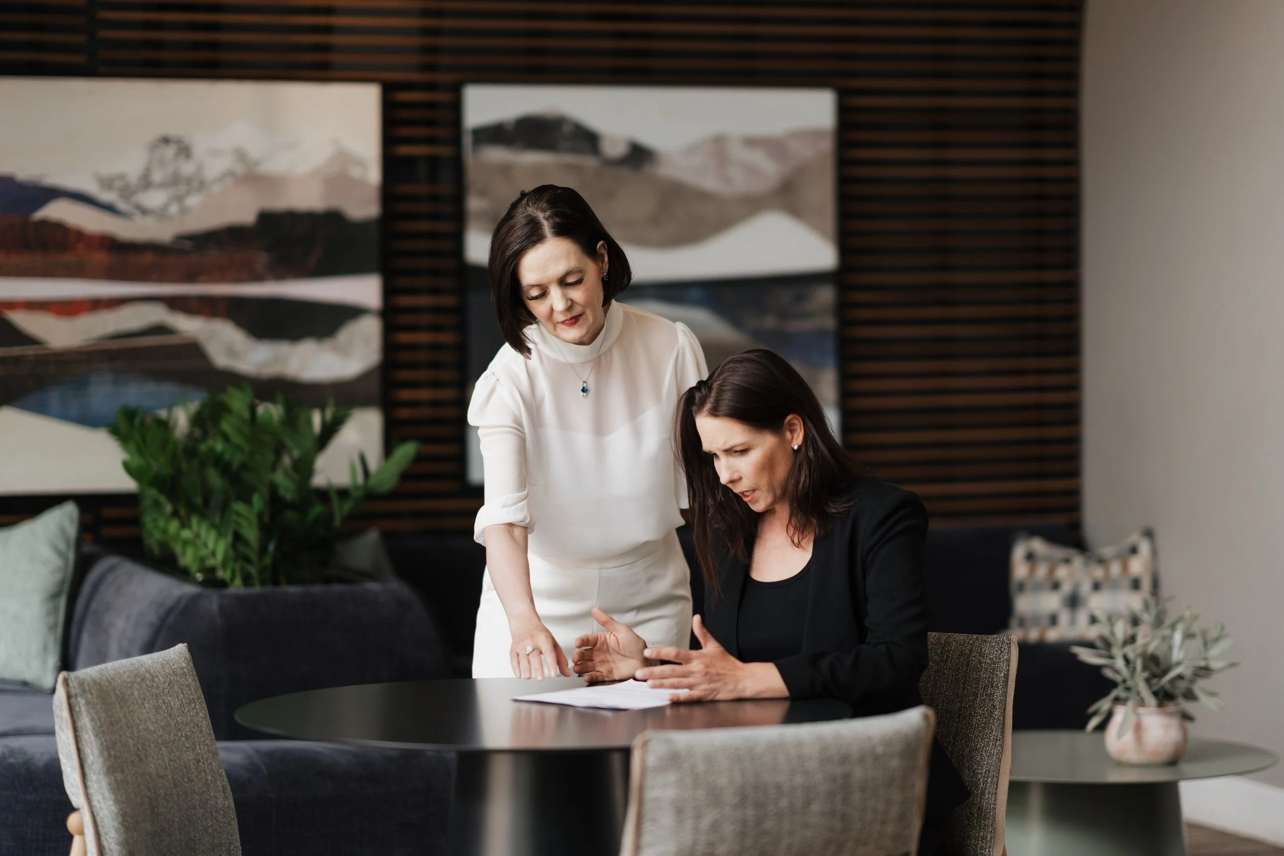 Two women engaged in a discussion at a dining table in a modern room. One woman standing and the other sitting, both looking at a device. Background features abstract mountain artwork and indoor plants.