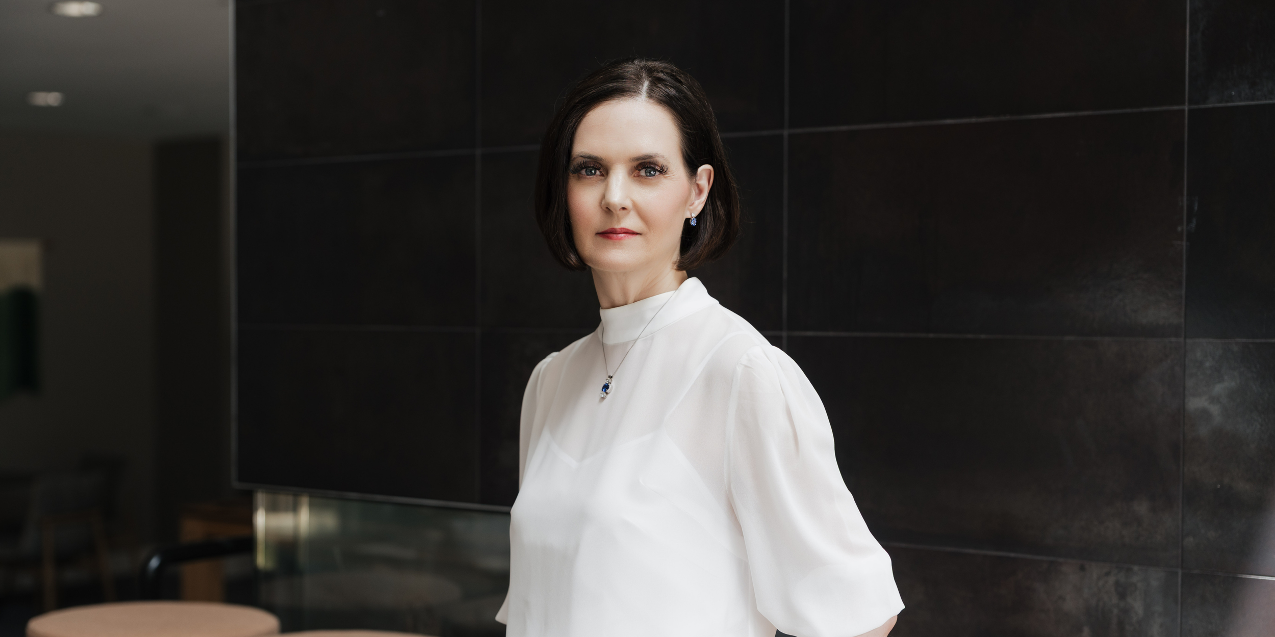 A woman with short dark hair and blue eyes wearing a white blouse and blue jewelry stands in front of a black tiled wall.