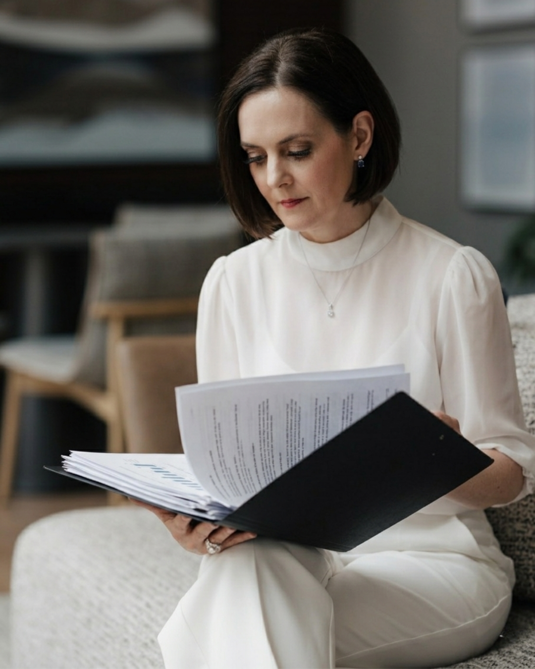 A woman with short dark hair and earrings reading a large black folder with papers inside, sitting on a light-colored chair in an indoor setting.