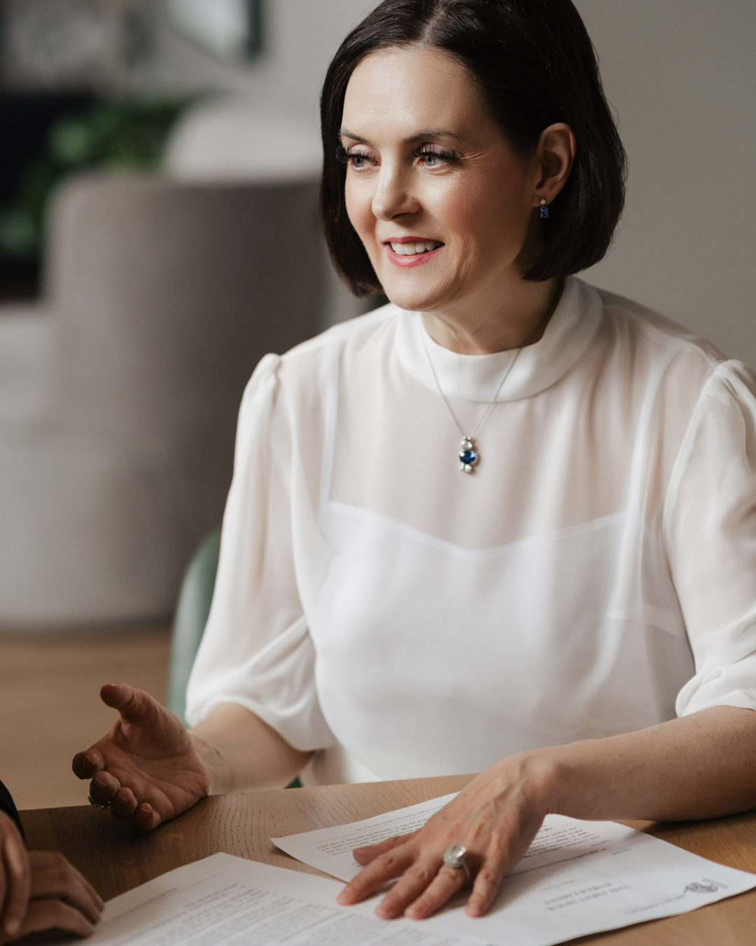 A woman with short dark hair, wearing a white blouse and jewelry, is sitting at a table with papers in front of her, smiling and talking to someone off-camera.