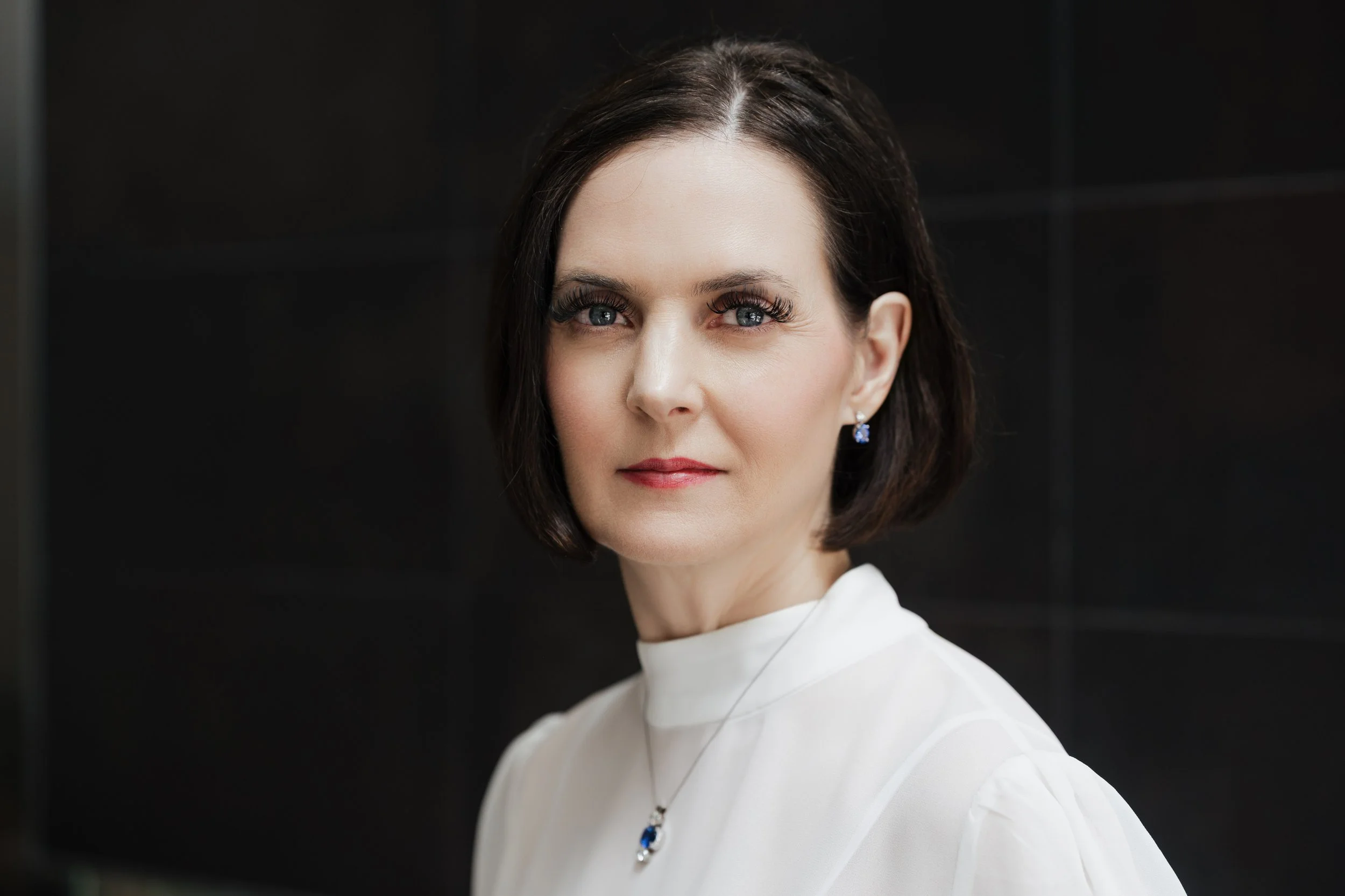 A woman with dark brown hair, light skin, blue eyes, wearing earrings and a white top, looking directly at the camera against a black background.