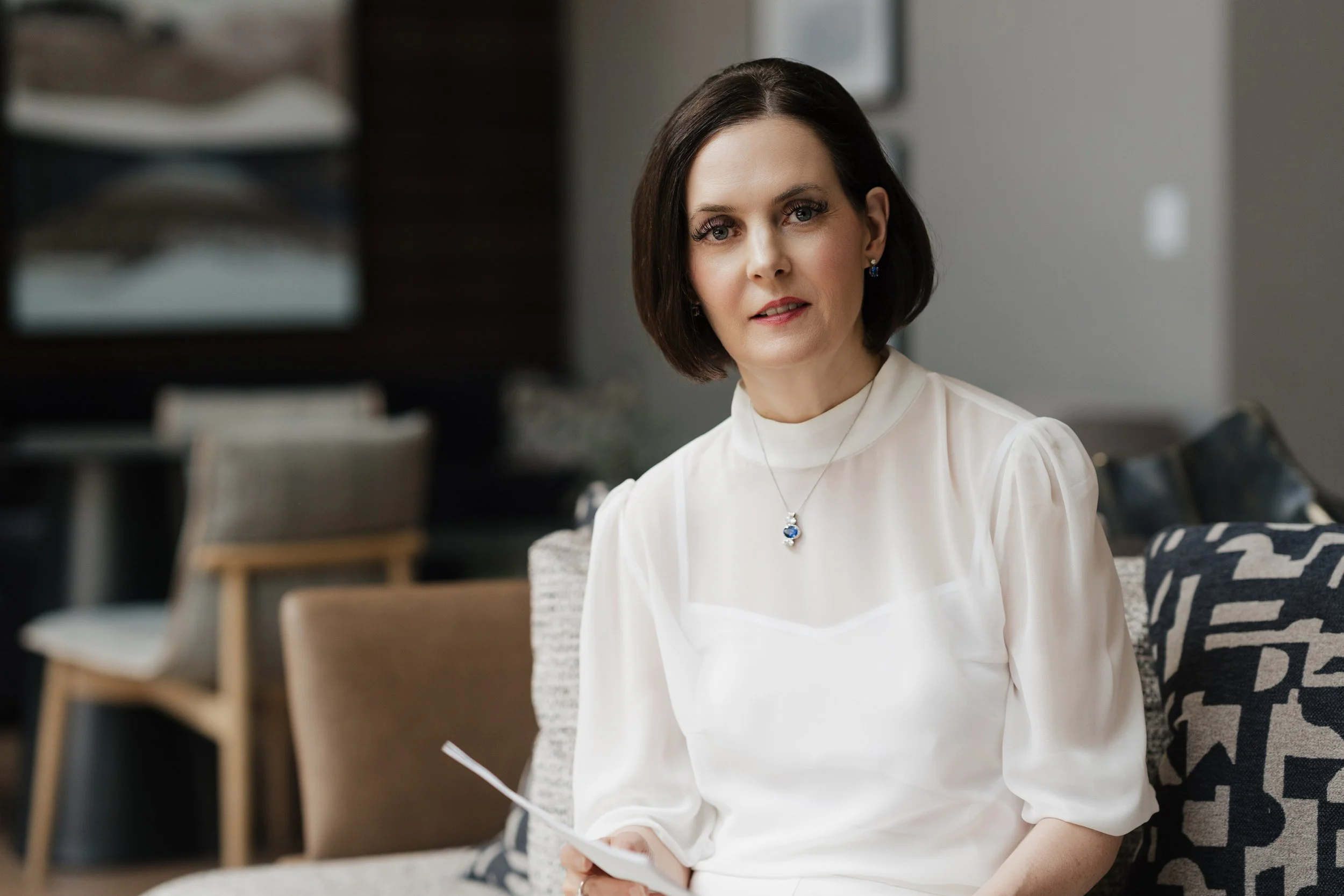 A woman with short dark hair, wearing a white blouse and blue jewelry, sitting on a patterned couch in a modern living room.