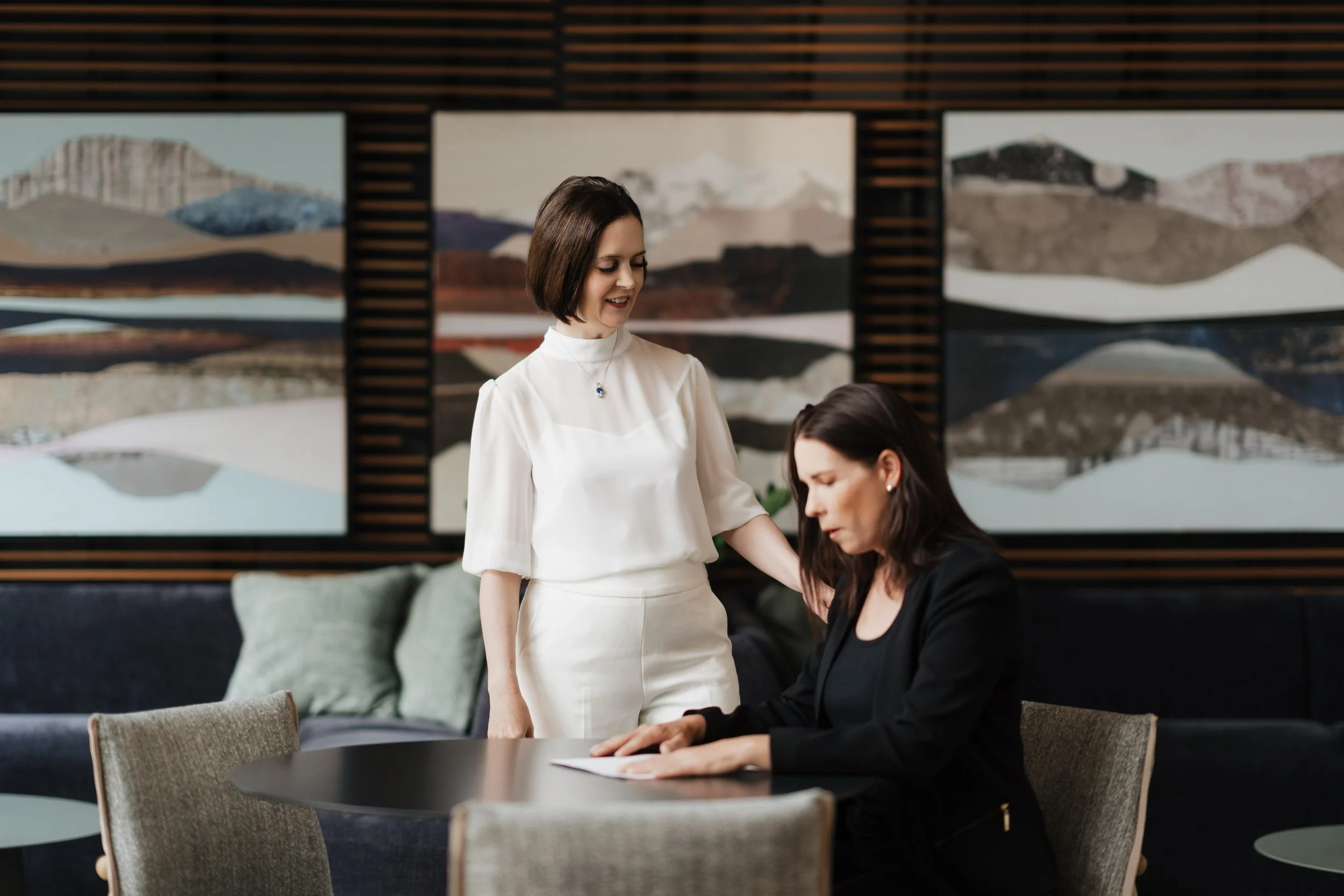 Two women in an art gallery, one standing and the other sitting at a table, with abstract landscape paintings on the wall behind them.