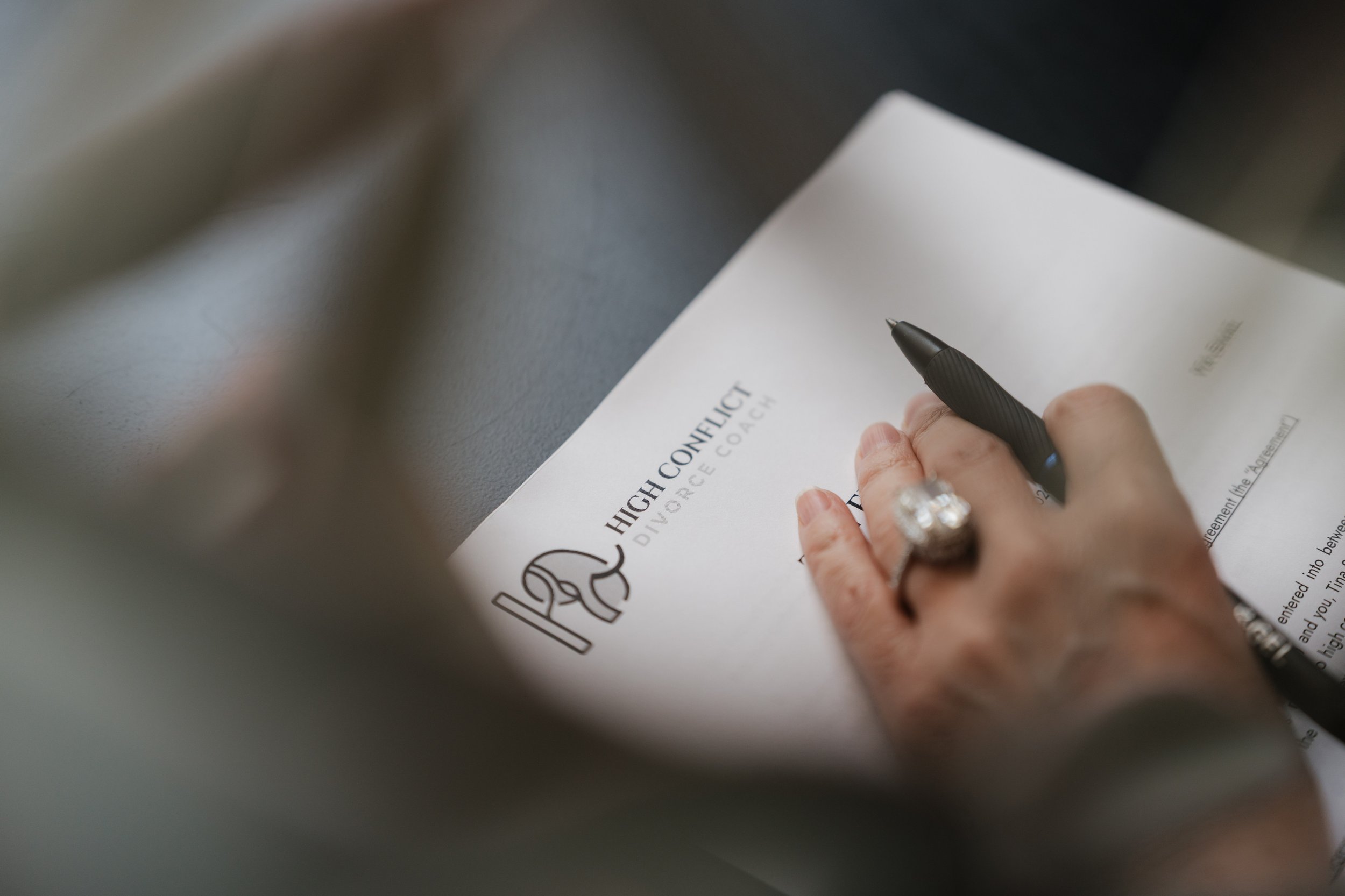 A person with a large ring on their finger is signing a document titled 'High Conflict Divorce Coach' with a black pen. The document is held down by their hand and is on a gray surface.