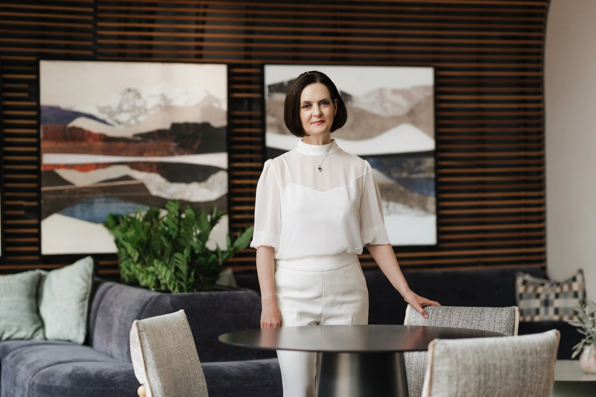 A woman with dark hair in a white outfit standing in a modern living room with abstract mountain artwork on the wall behind her and a gray sofa with green cushions in the foreground.