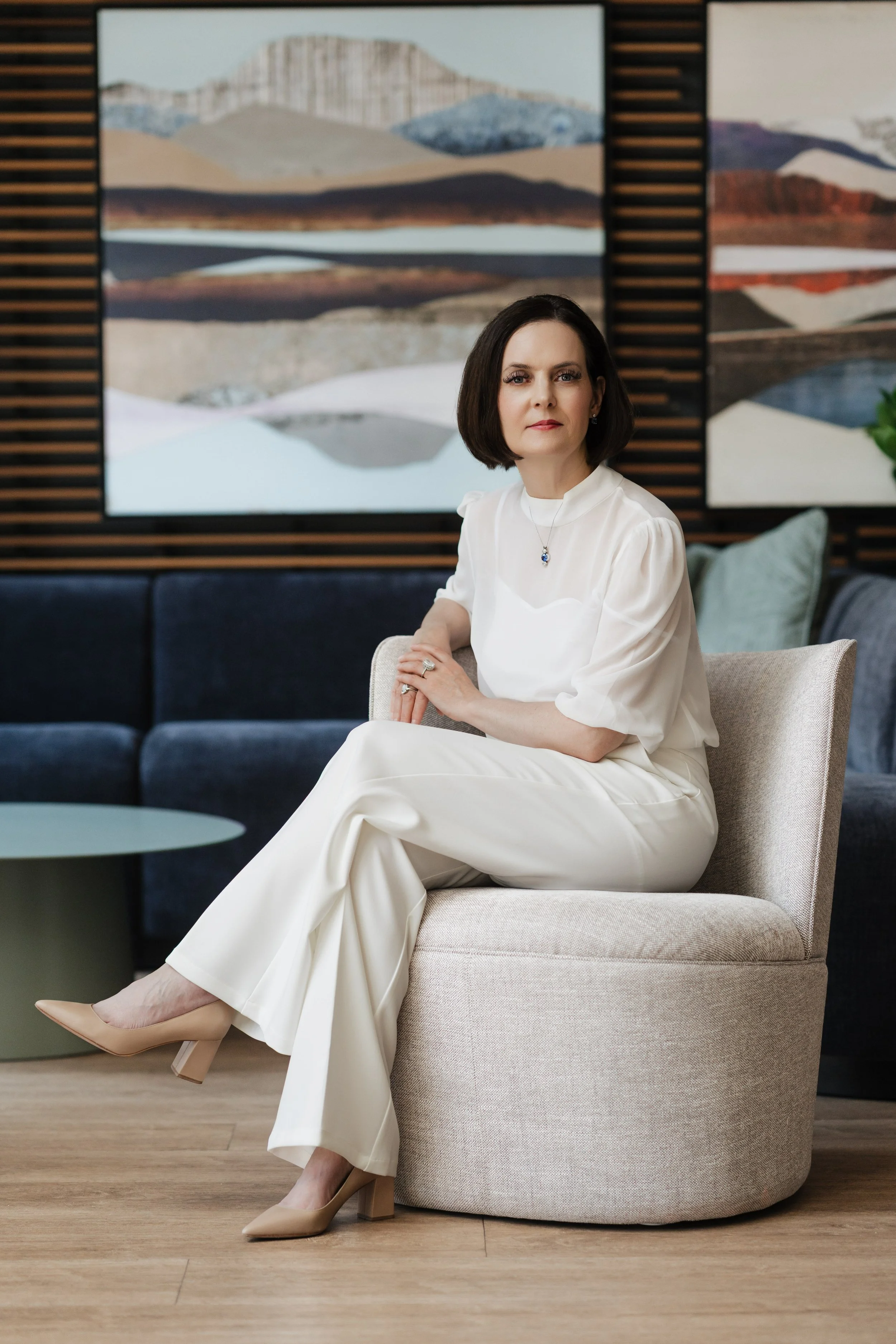 A woman sitting on a beige chair in a modern interior with abstract landscape paintings on the wall behind her, dressed in a white outfit with beige heels.