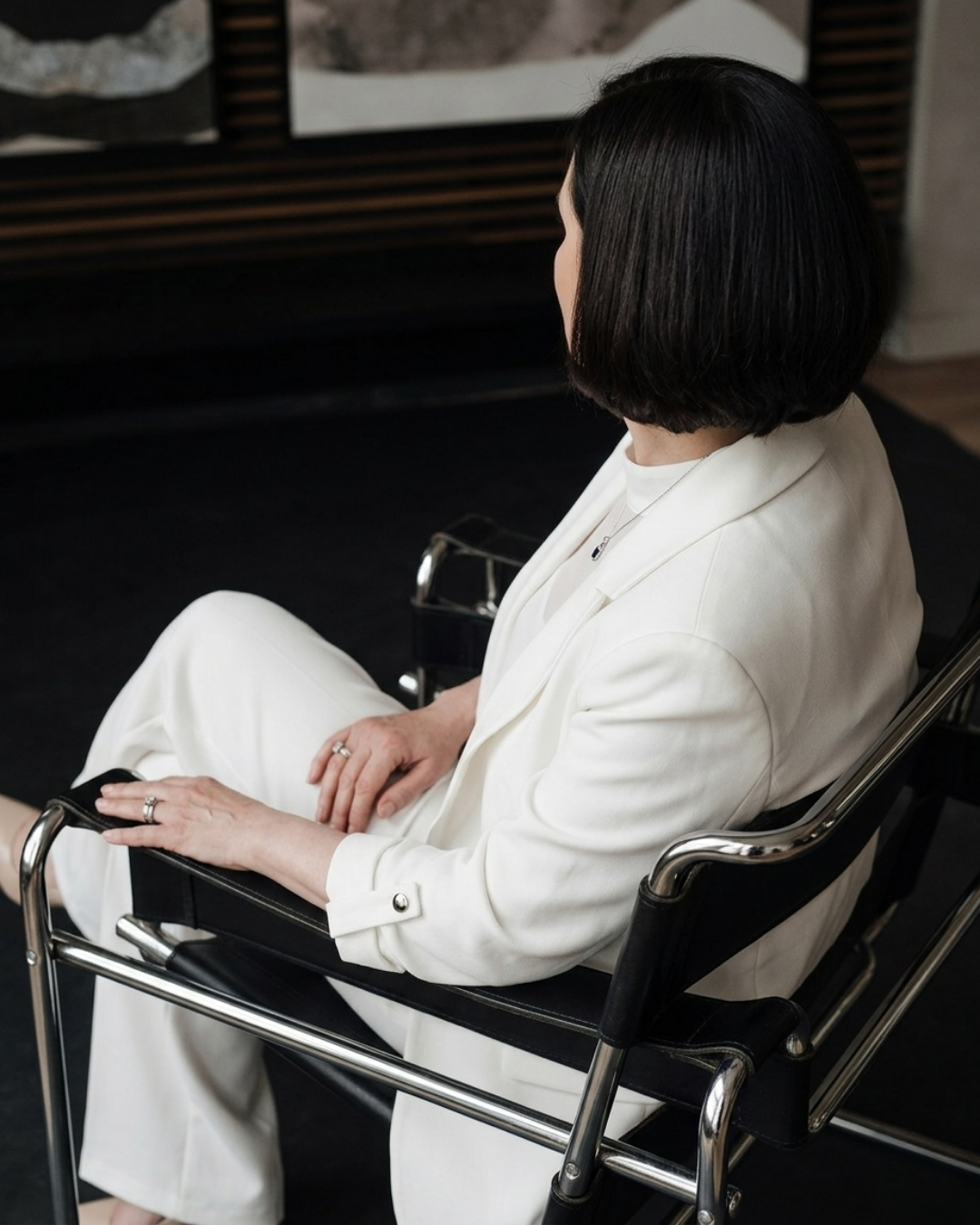 A woman with black hair wearing a white suit sitting in a black and chrome chair, looking to the side, with paintings on the wall behind her.