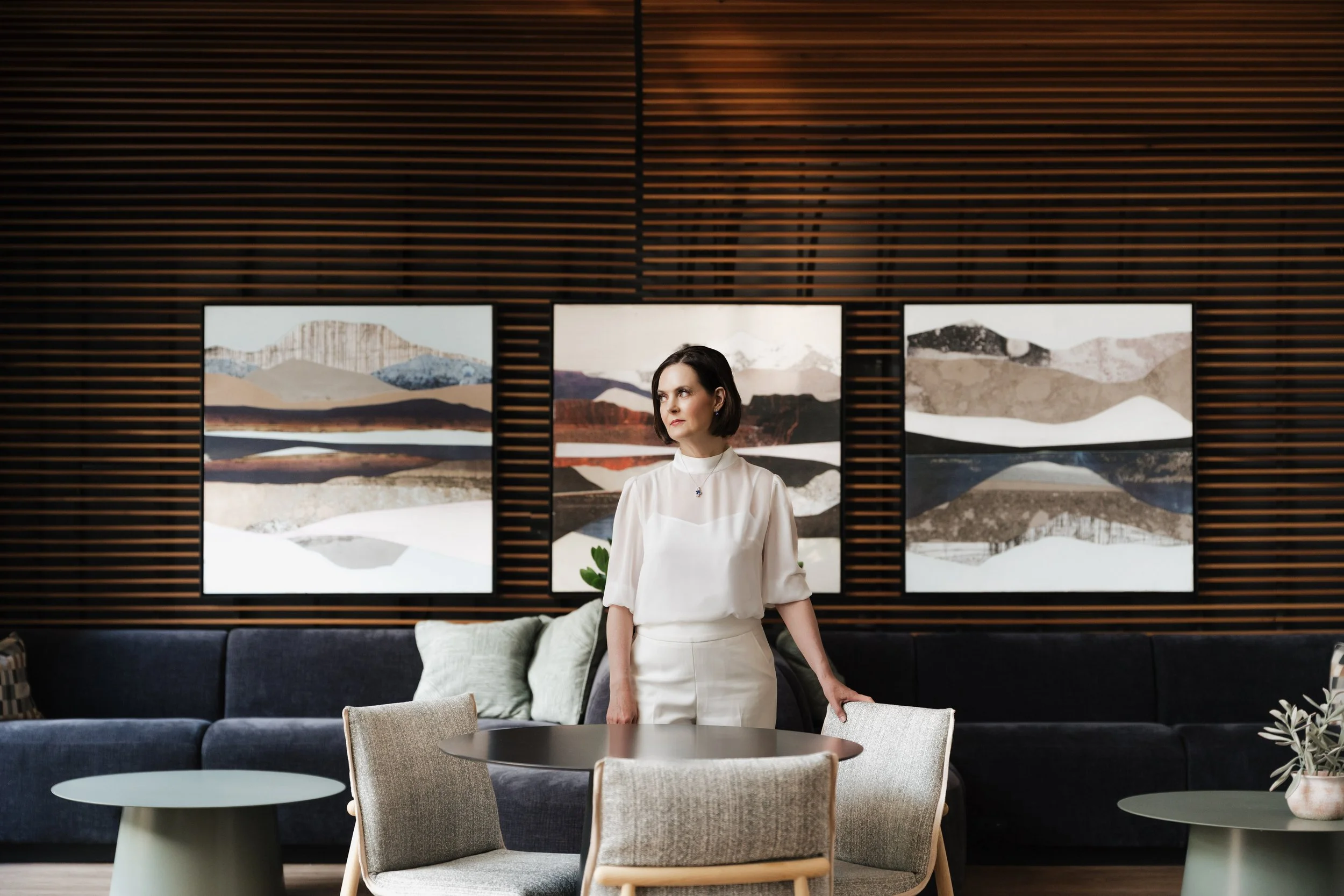 A woman in a white outfit standing in a modern living room with three landscape artwork pieces on a dark wood-paneled wall behind her.