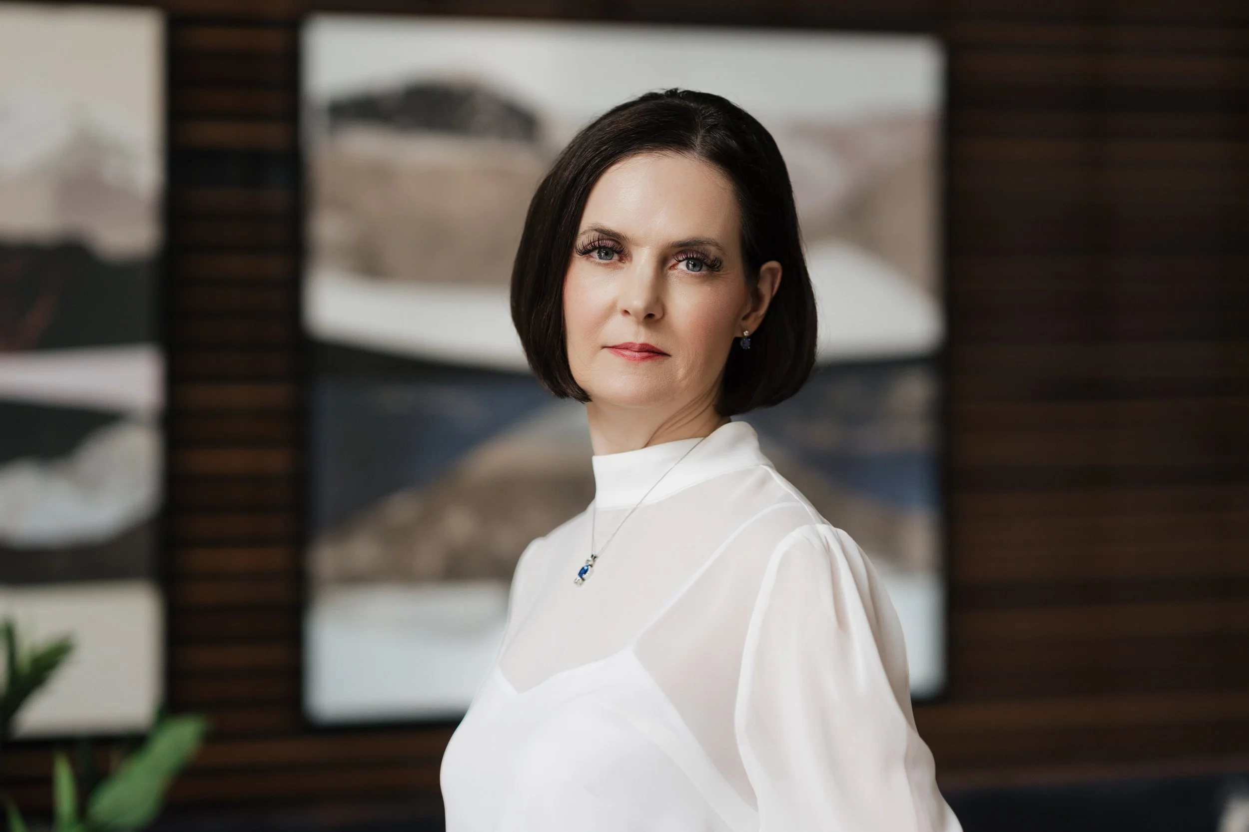 A woman with dark hair, wearing a white dress and jewelry, posing in front of blurred background of artwork and dark wood wall panels.