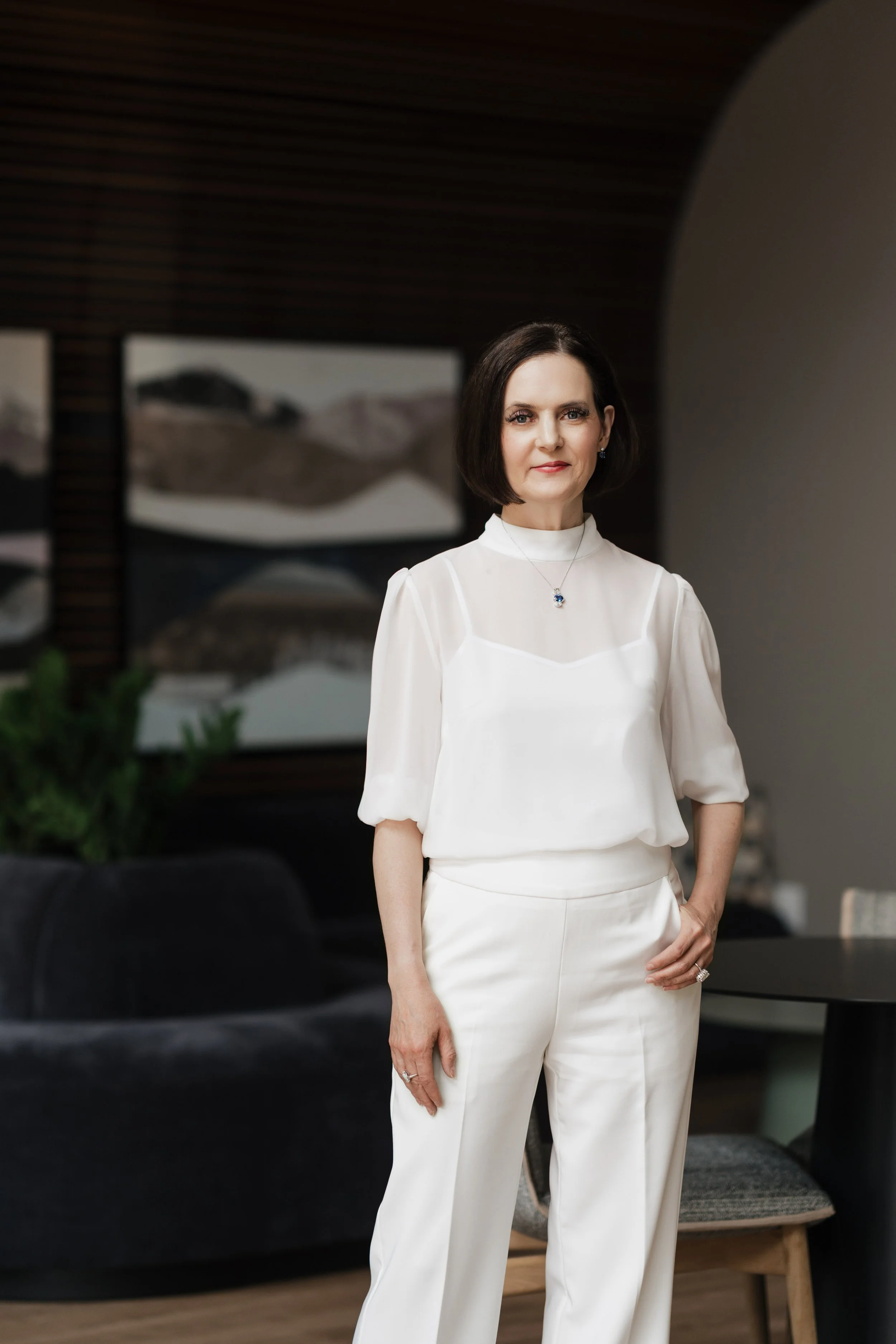 A woman with dark, shoulder-length hair wearing a white blouse with puffed sleeves and high-waisted white trousers, standing inside a modern room with wooden wall accents and abstract artwork in the background.