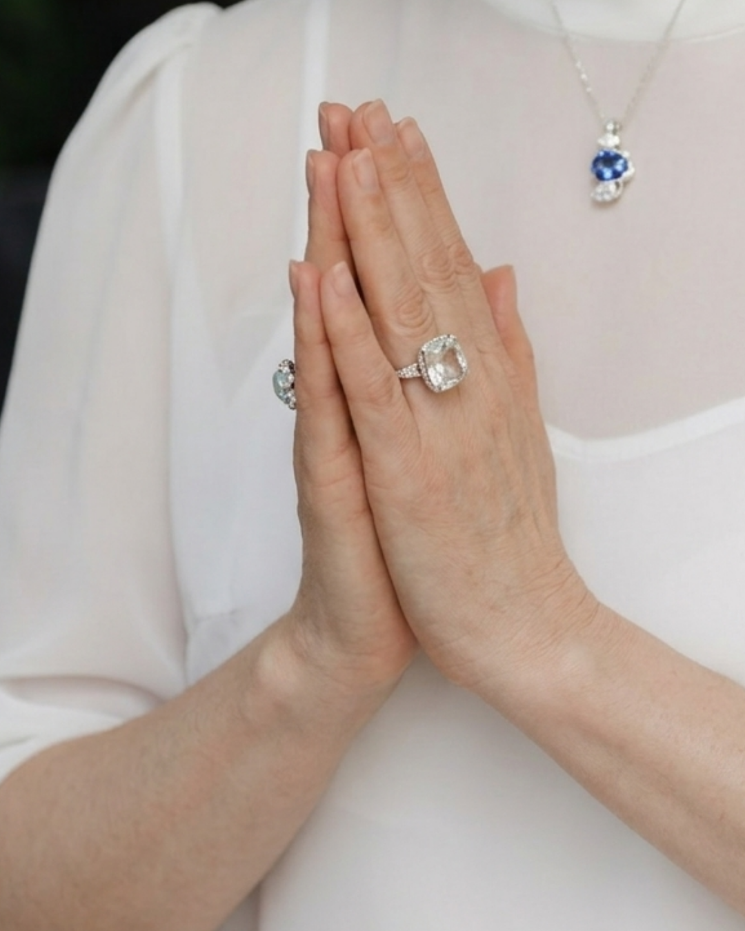 Person with hands together, wearing diamond rings and a necklace with blue and white stones, on a white top.