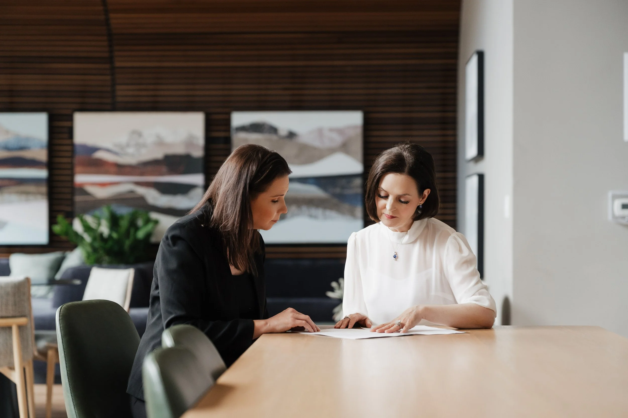 Two women are sitting at a table, reviewing a document together in a modern, well-lit office space.
