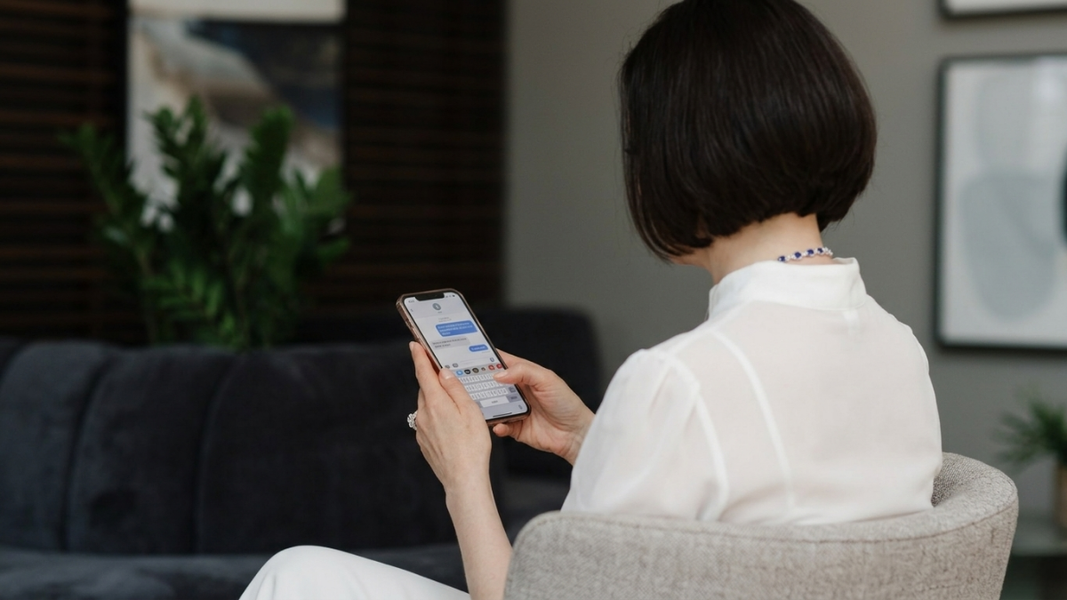 A woman with short dark hair reading a text message on her smartphone while sitting in a modern living room.