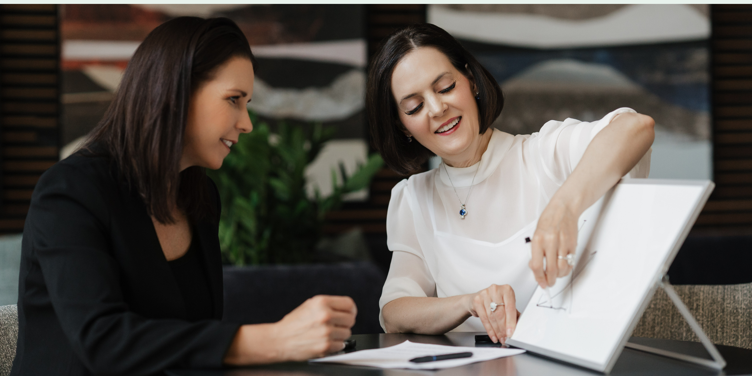 Two women are sitting at a table looking at a tablet, smiling, with documents and a pen on the table.