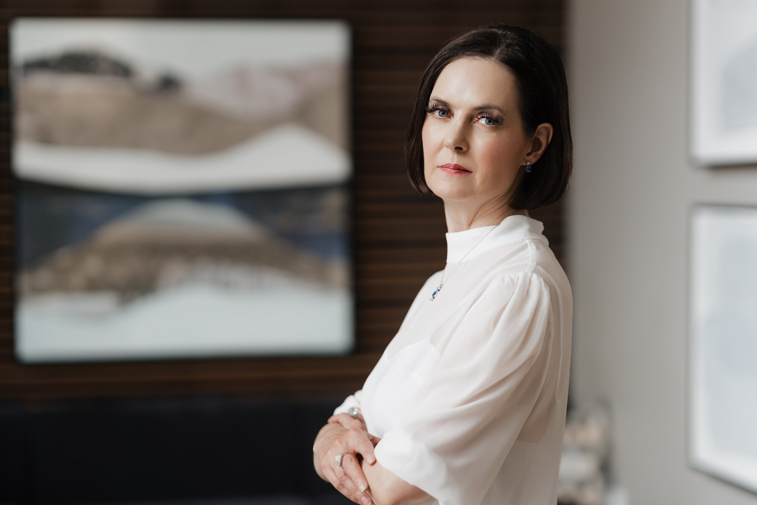 A woman with short dark hair and blue eyes standing near a piano, wearing a white blouse and earrings, looking at the camera.