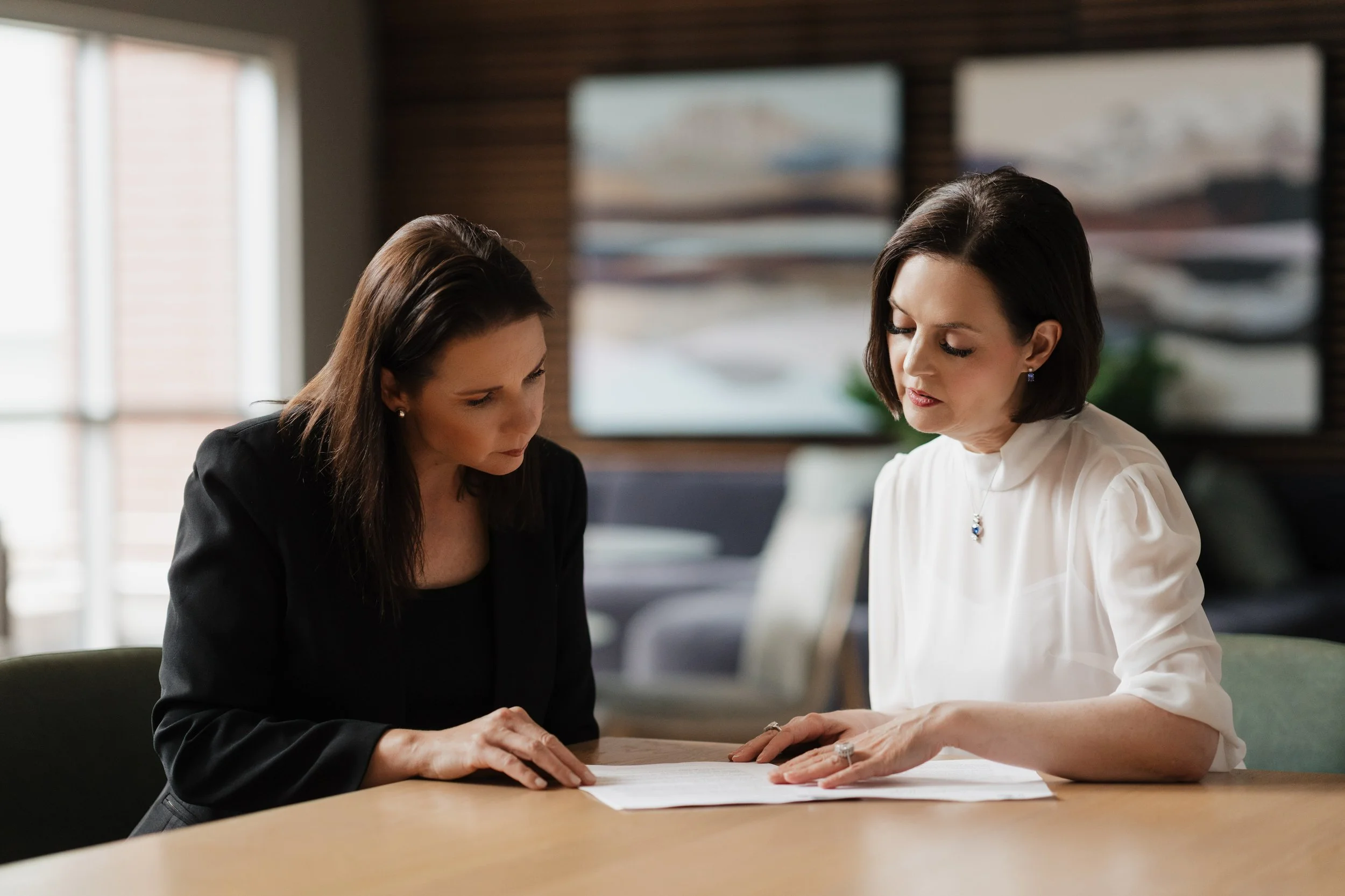 Two women sitting at a table, holding hands, with eyes closed and heads bowed, in a professional or serious setting.