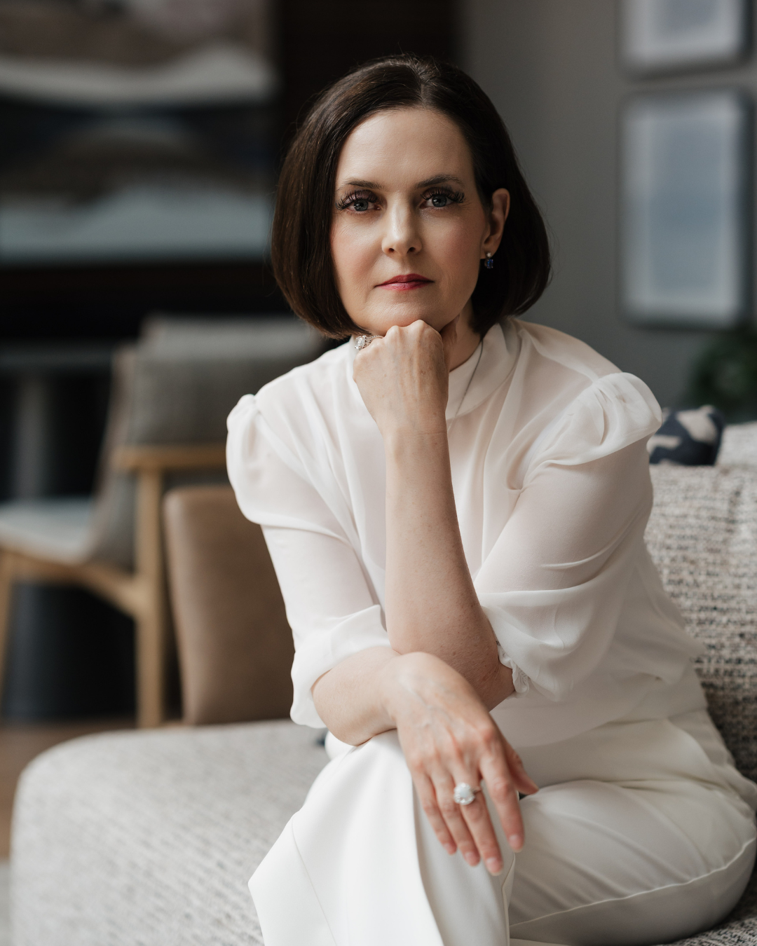 A woman with short dark hair and blue eyes sitting on a gray couch, wearing a white dress, with her chin resting on her hand, posing confidently in a modern interior.