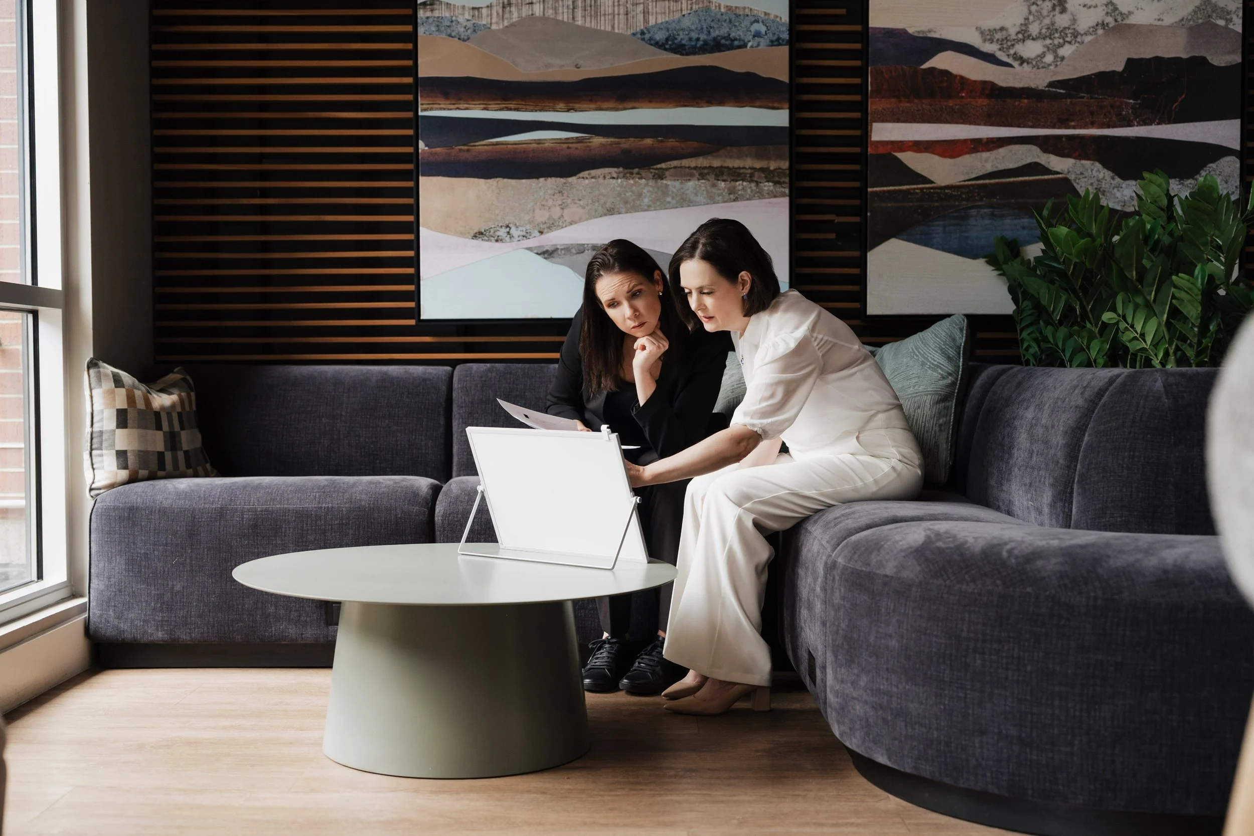 Two women sitting on a dark gray couch, looking at documents together. They are in a modern living room with abstract landscape artwork on the wall and a large potted plant beside them. There is a round coffee table with a white display stand in front of them.