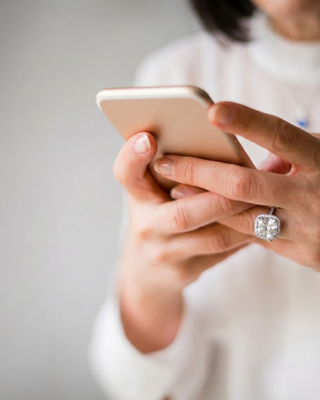 A woman with a diamond ring on her finger is holding and looking at her smartphone, with a light-colored blurred background.