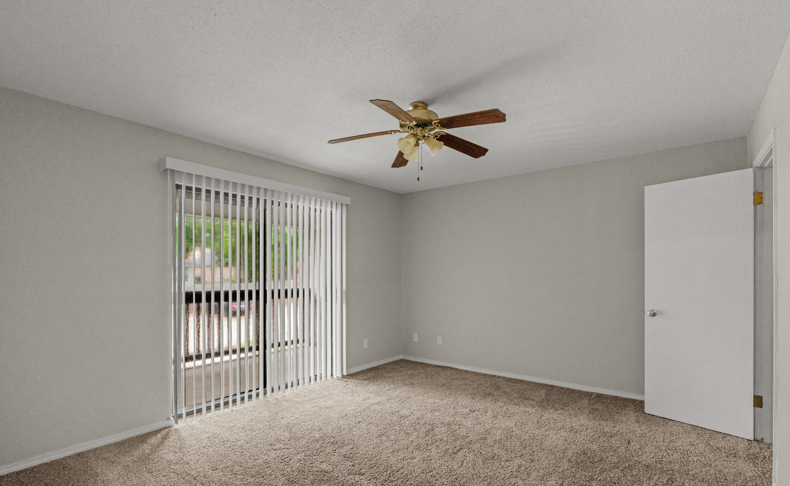 Empty living room with beige carpet, white walls, sliding glass door with vertical blinds, open door, ceiling fan with wooden blades.