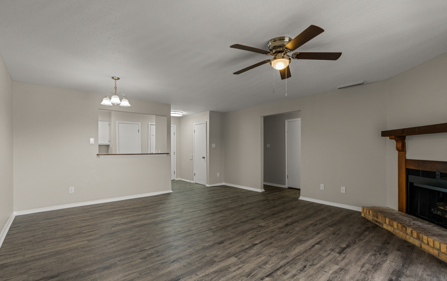 Empty living room with wood flooring, ceiling fan, fireplace with wood mantel, and hallway leading to other rooms.