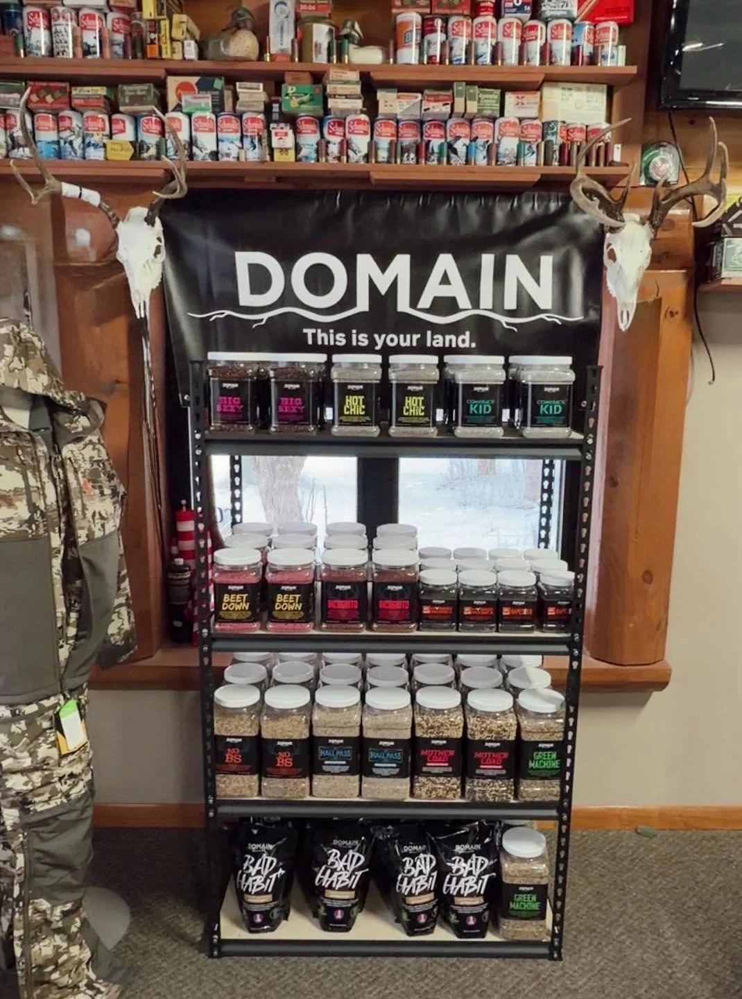 A display shelf with various jars of spices and seasonings, with a black banner labeled 'DOMAIN' and 'This is your land.' on top, flanked by deer skulls with antlers, in a rustic store setting.
