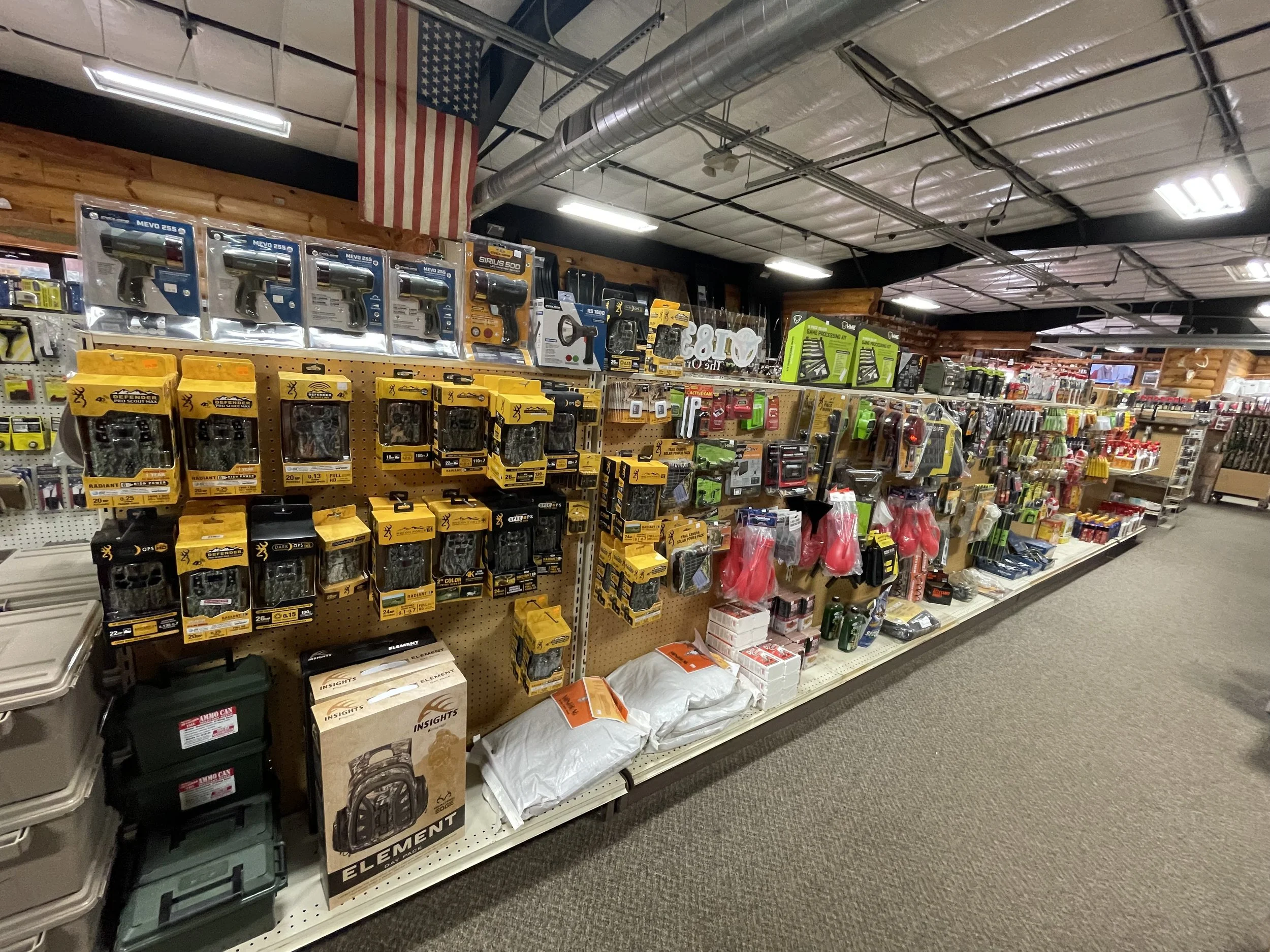 Hardware store aisle with various tools and supplies, including spray paint, spray guns, and safety gear, underneath a hanging American flag.