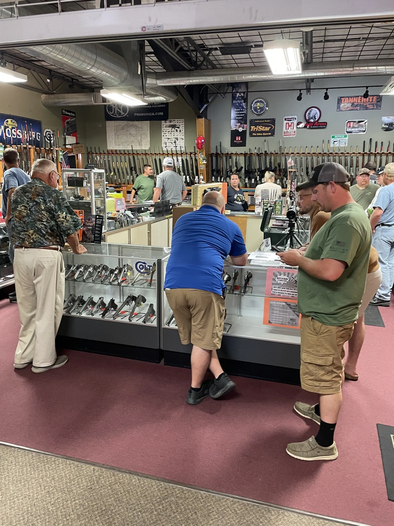 People shopping for firearms at a gun store, with rifles displayed on the wall behind the counter.
