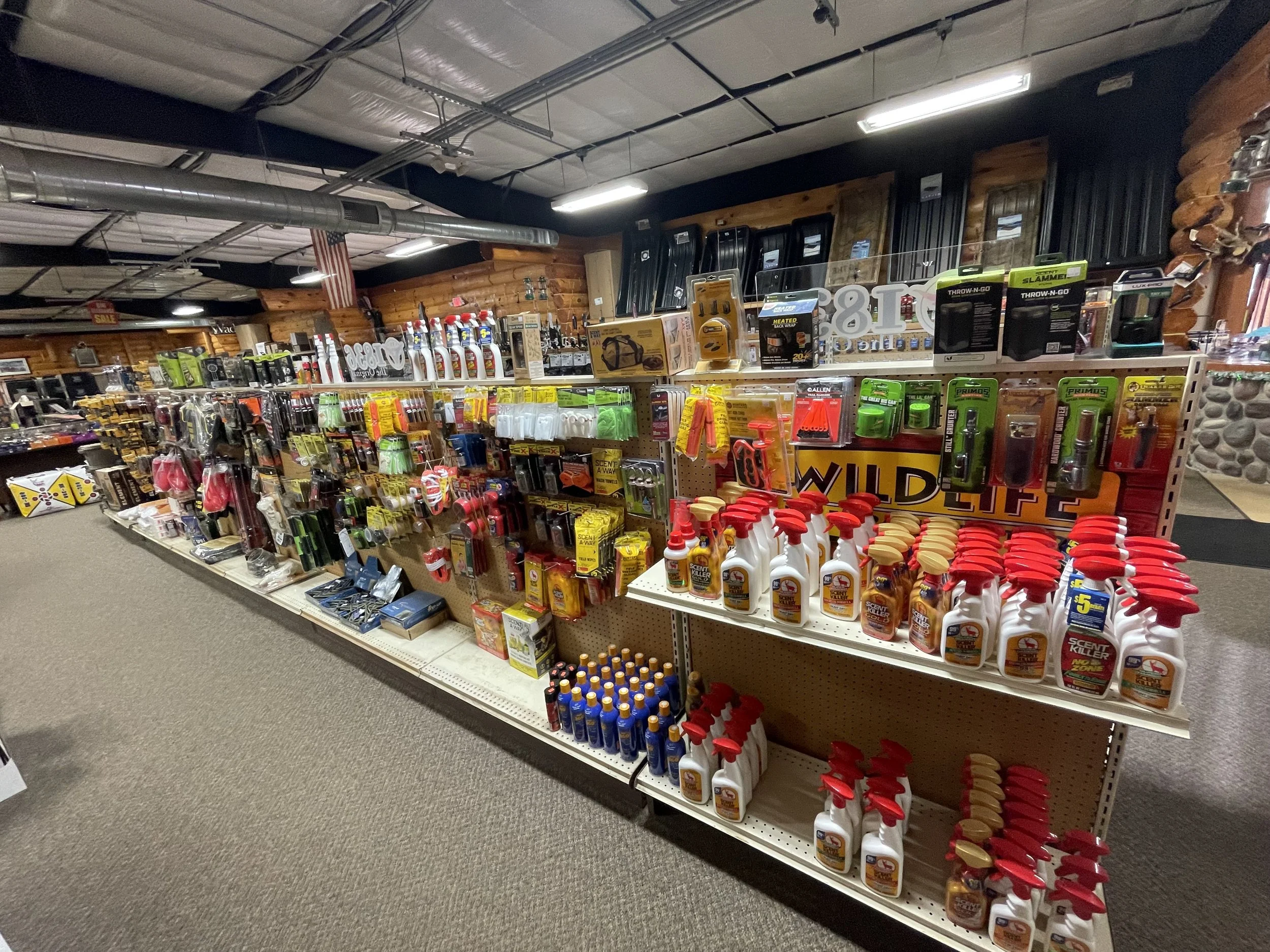 A store aisle with shelves stocked with various car care products, cleaning supplies, and tools. The background includes a log cabin-style interior with wooden walls and an American flag hanging from the ceiling.