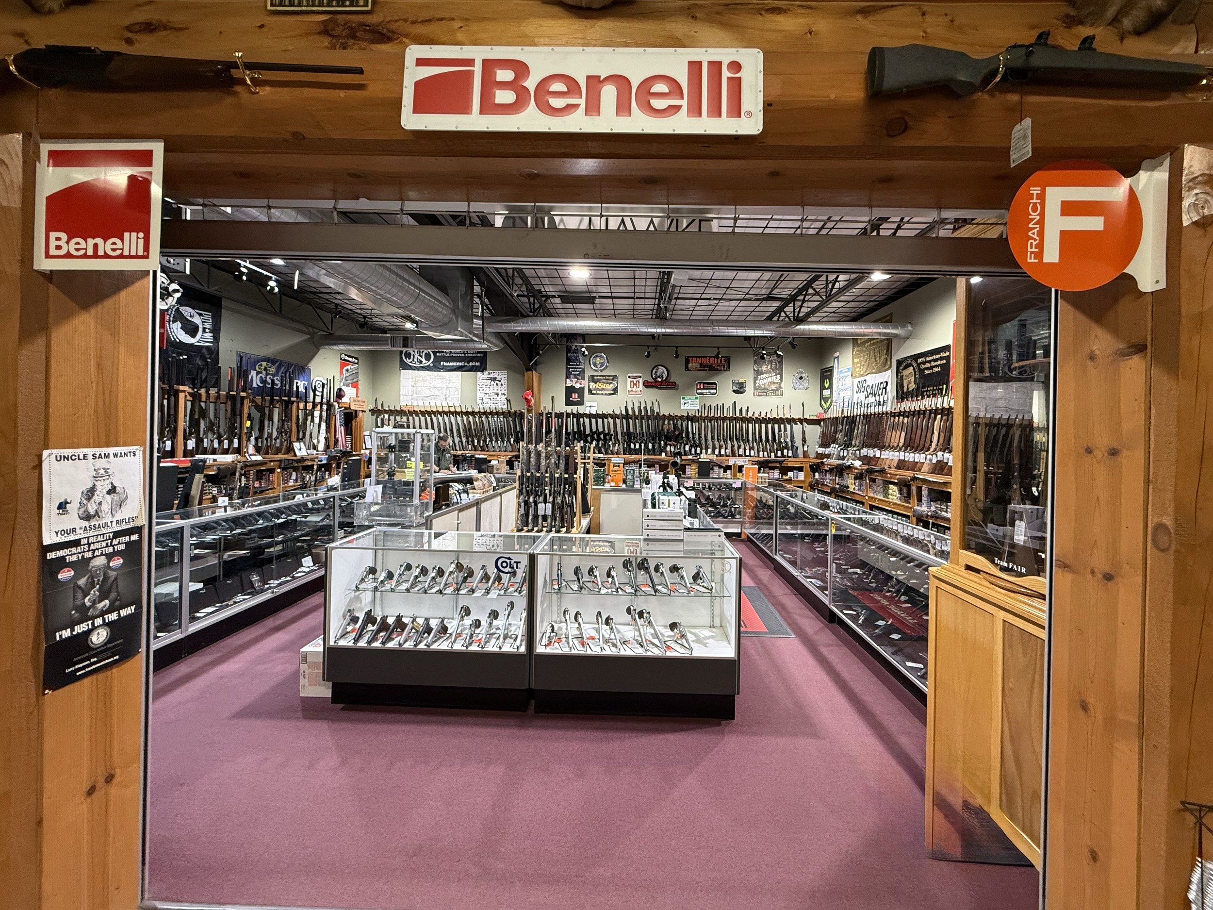 View of a firearm store with rifles displayed on racks and posters on the walls, seen through a wooden doorway with a 'Benelli' sign above.