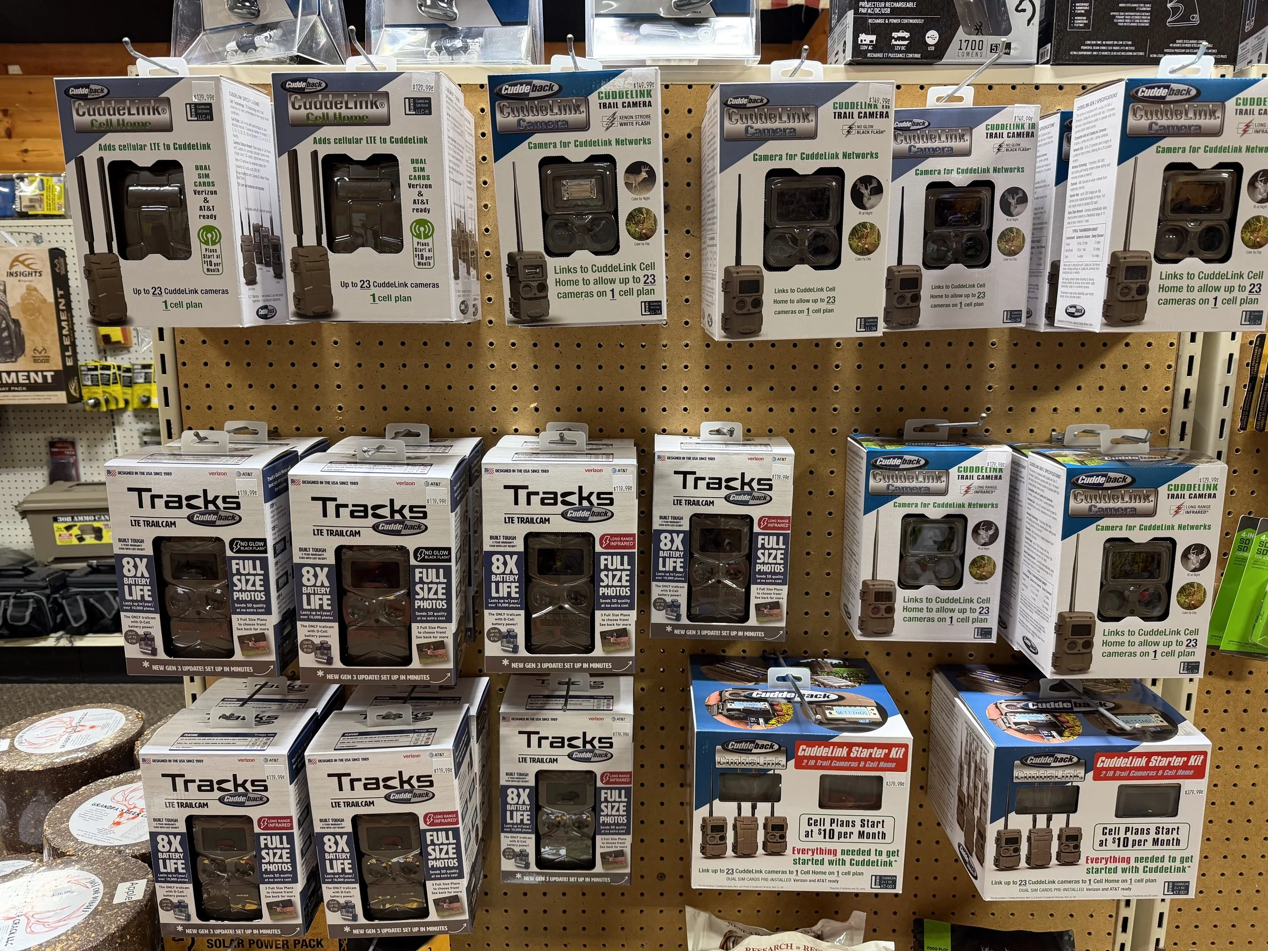 Display of CuddeLink security cameras and trail cameras in packaging on store shelf, featuring models with cell plans, long battery life, and infrared night vision.