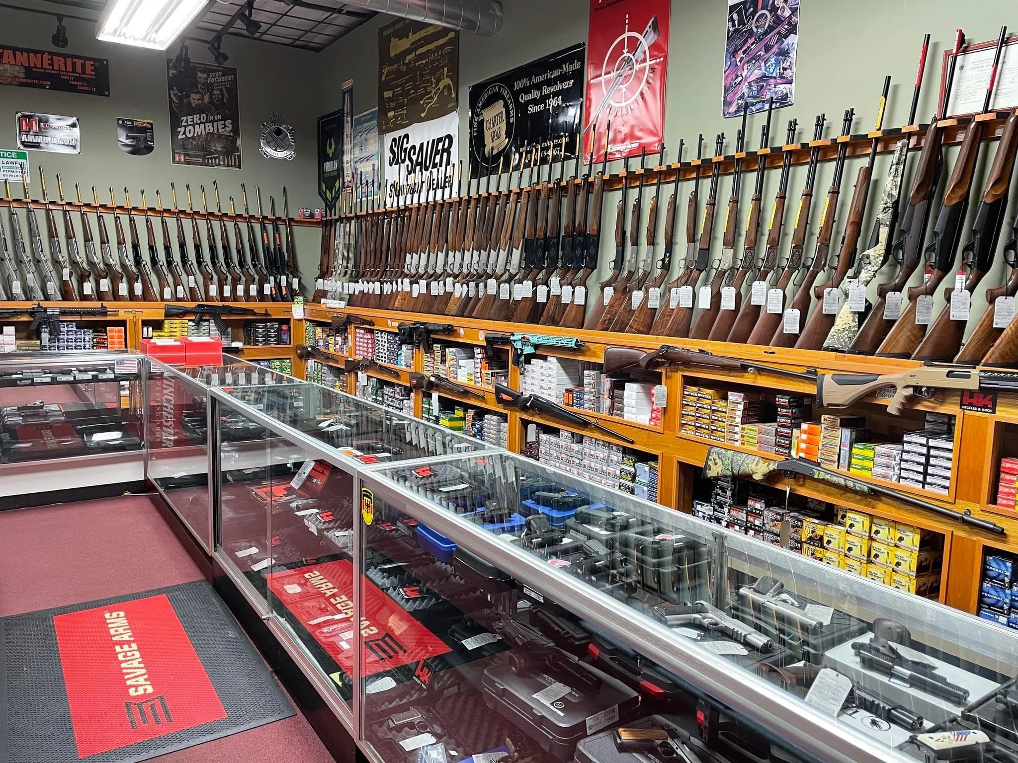 Store aisle with rifles displayed on a wall, along with gunpowder and ammo in glass cases below. Posters and banners related to firearms on the wall.