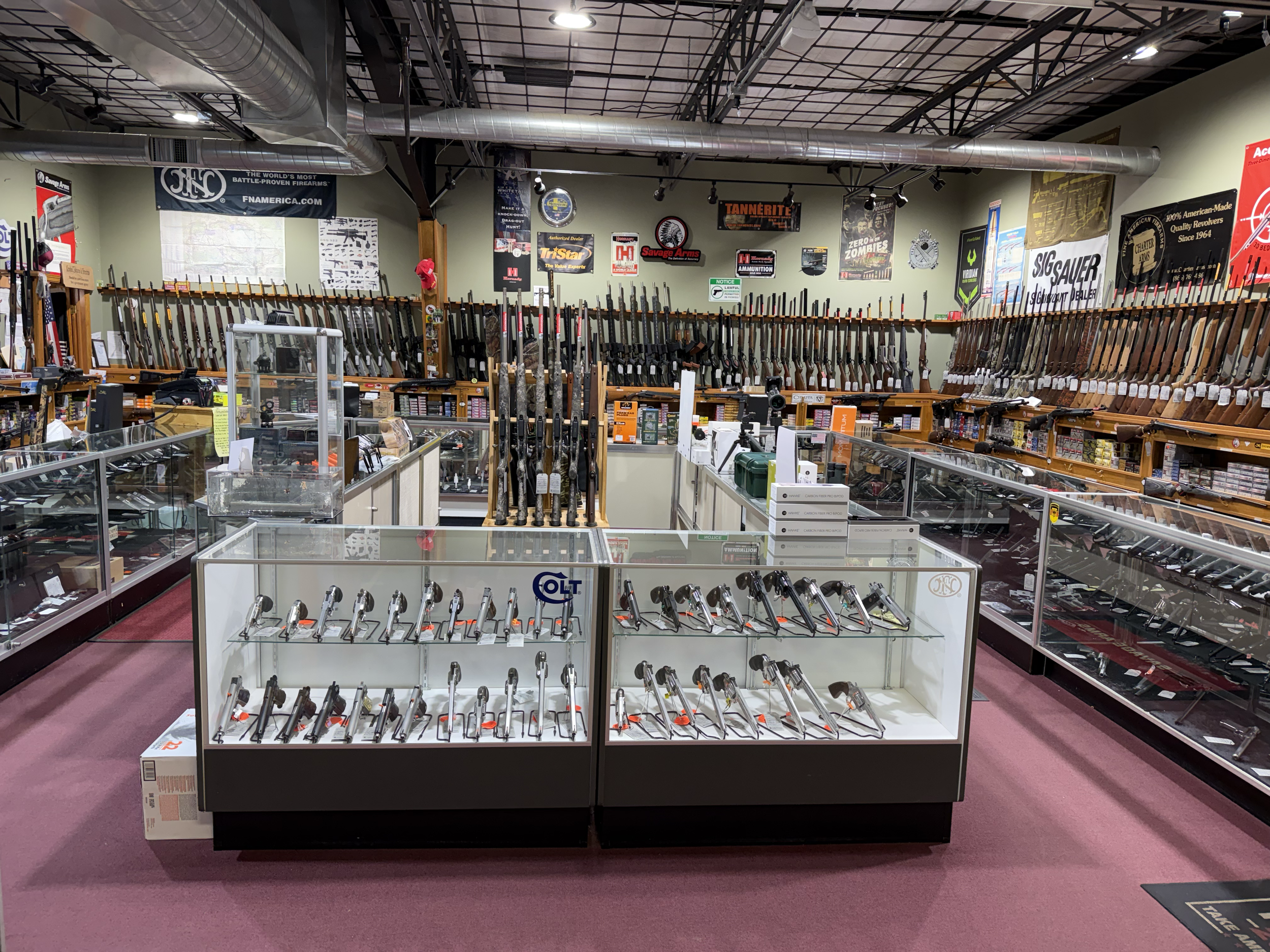 A display of firearms inside a store, with rifles and handguns arranged on racks and in glass cases, and various firearm brand and advertisement signs on the walls.