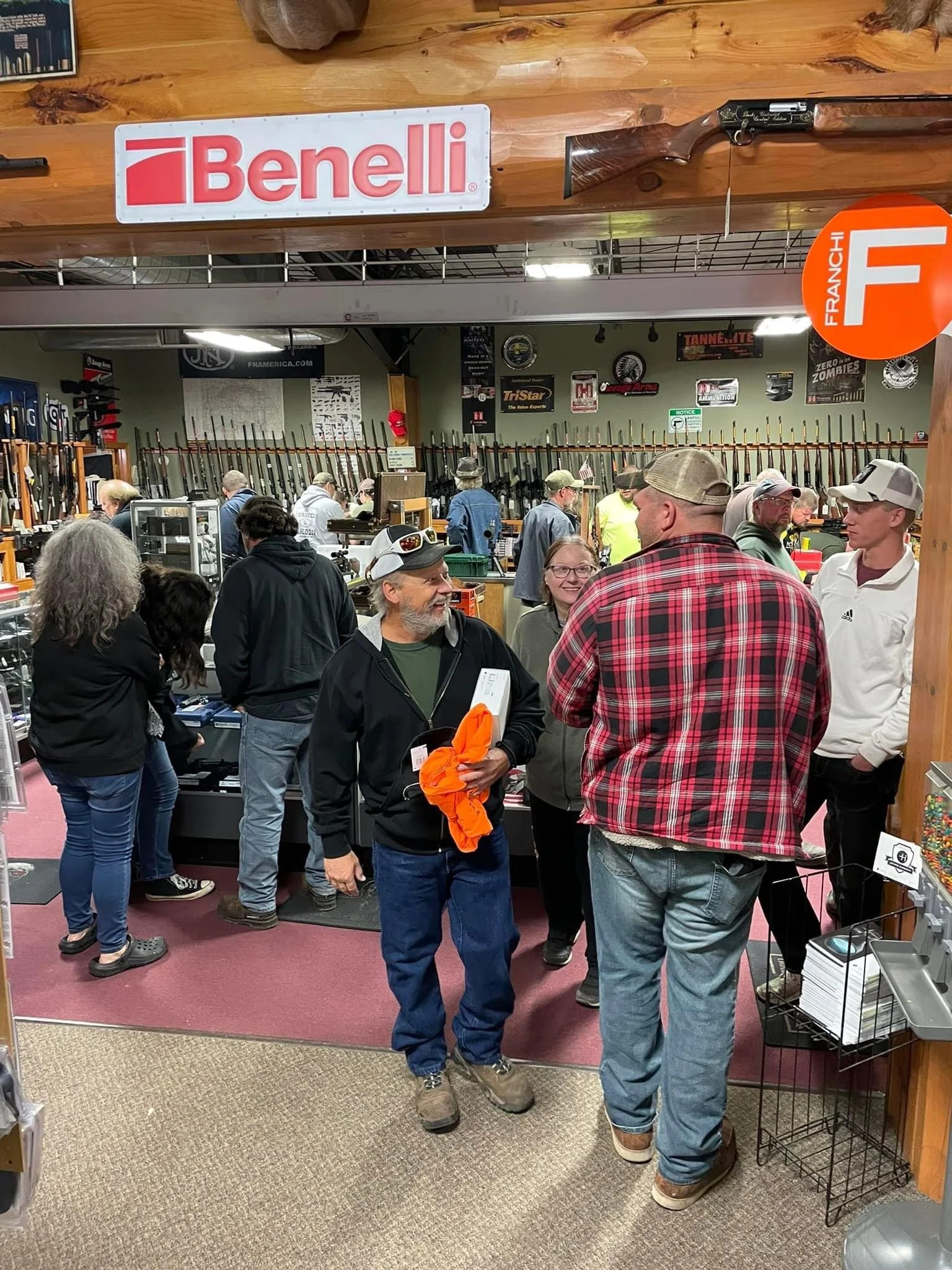 People shopping for firearms at a gun store, with rifles displayed on the wall in the background, and a Benelli sign hanging from the ceiling.
