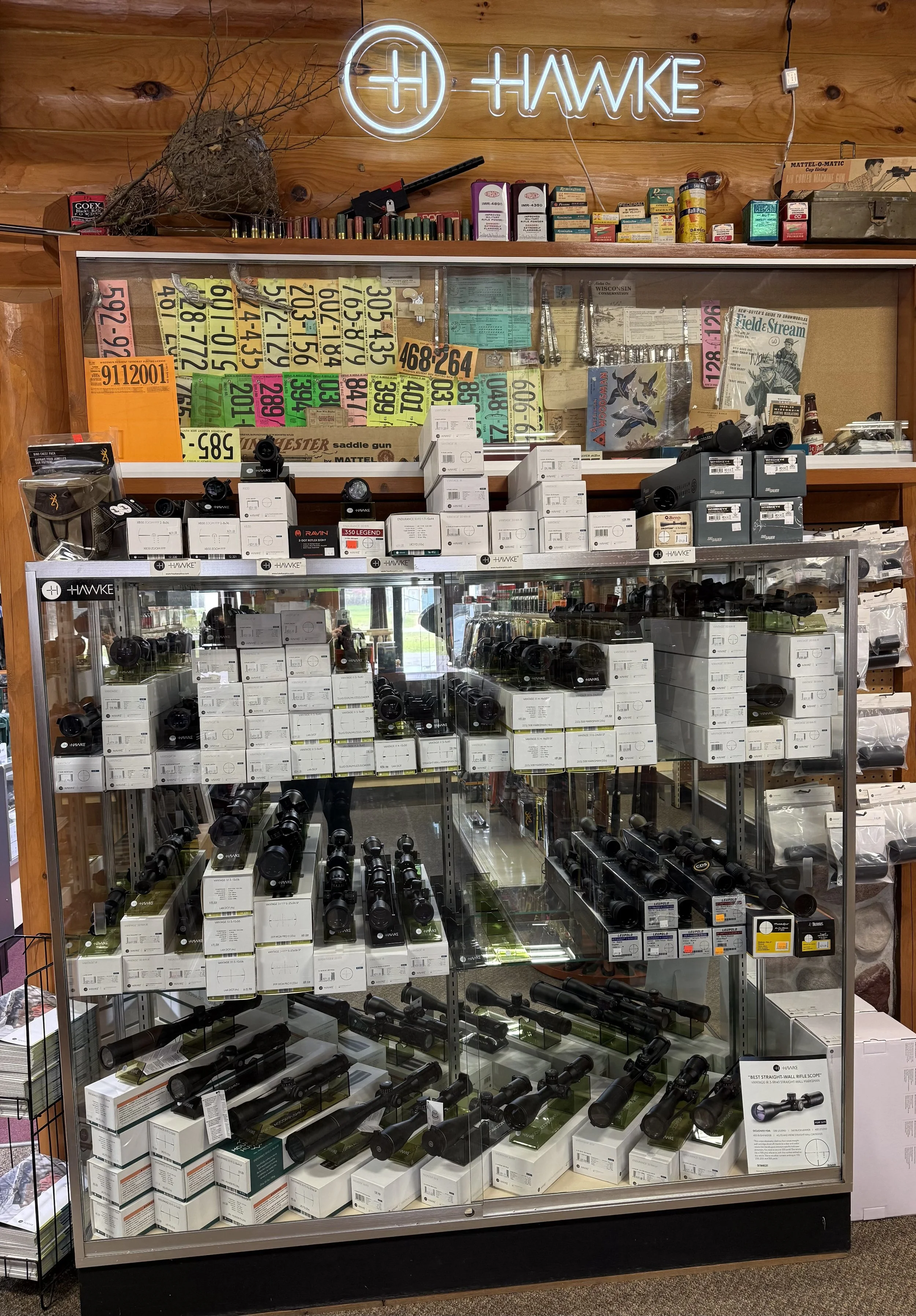 Display case filled with scopes and related accessories in a store, with a wooden wall and neon sign that says 'Hawke' in the background.