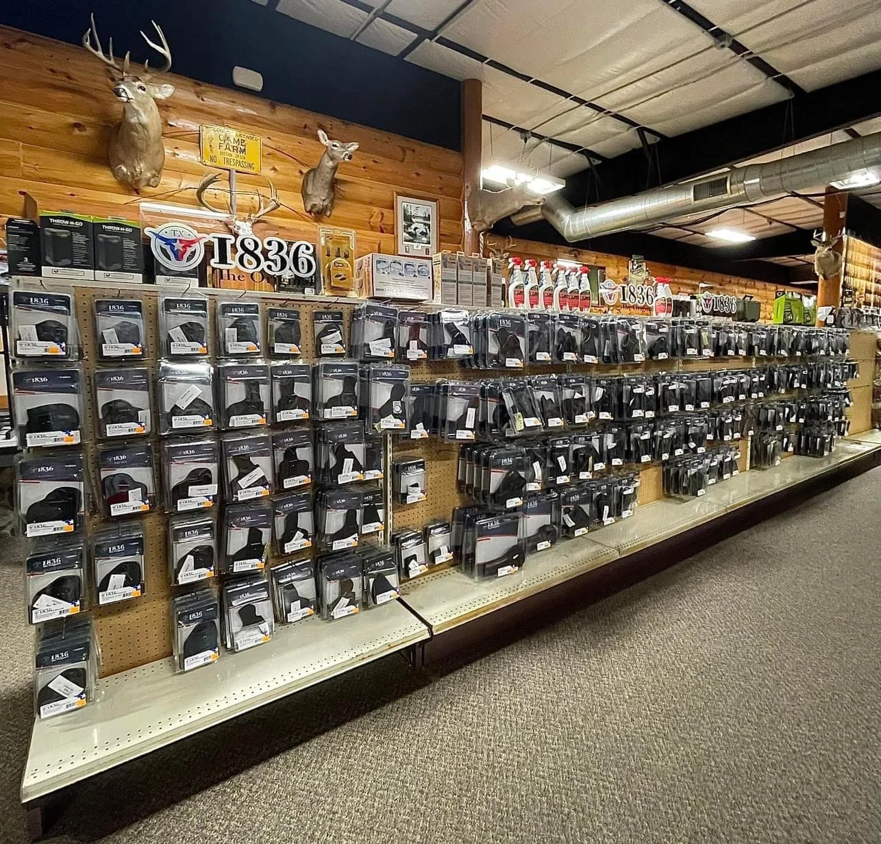 Store aisle with gun accessories in packages on shelves, parks on a wooden wall, deer heads mounted on wall, and signs with the text '1836' and 'Game Farm'.