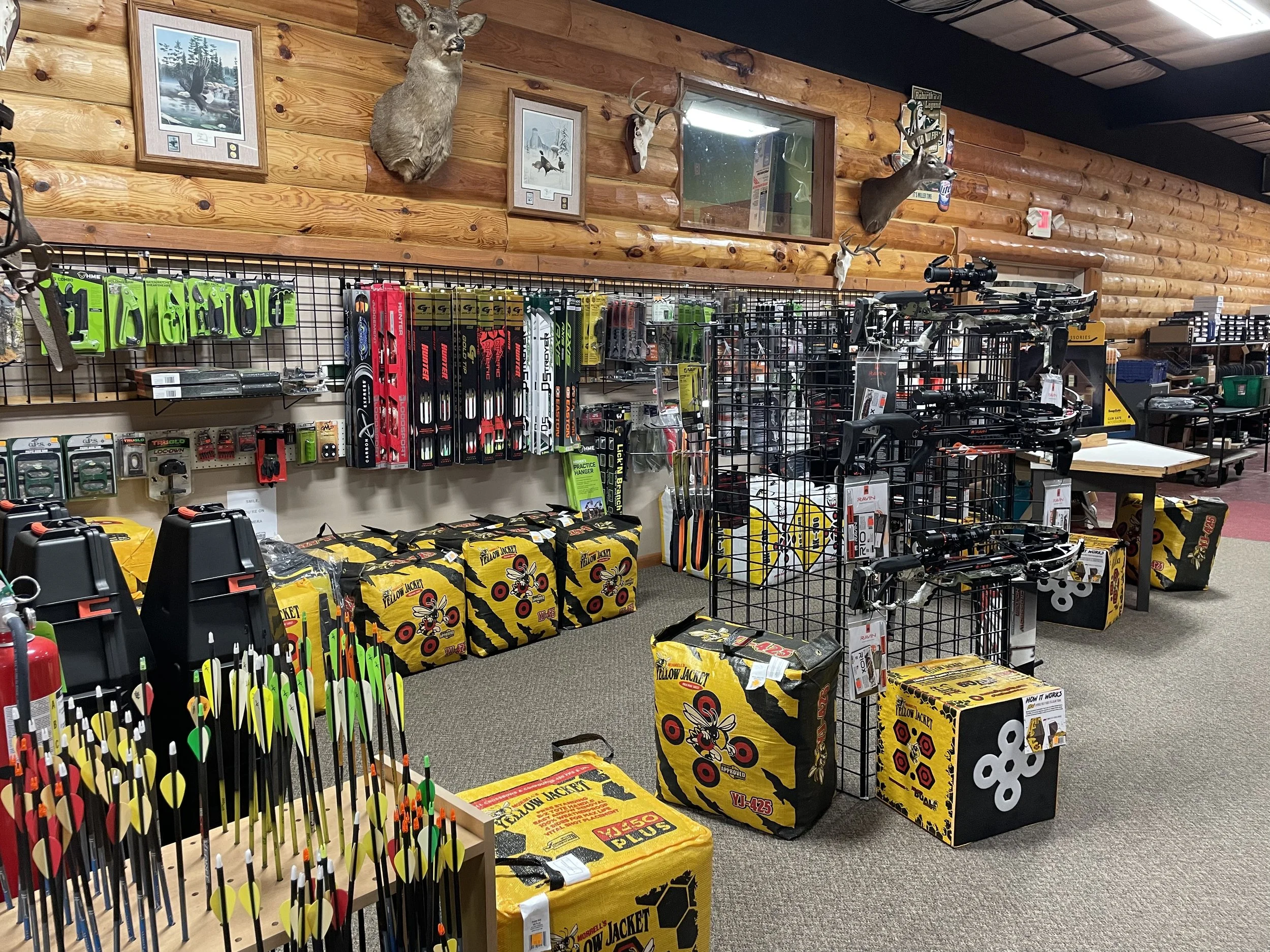 Interior of a sporting goods store with archery supplies, including colorful arrows and boxes of archery targets, with wood-paneled walls decorated with mounted deer and elk heads.