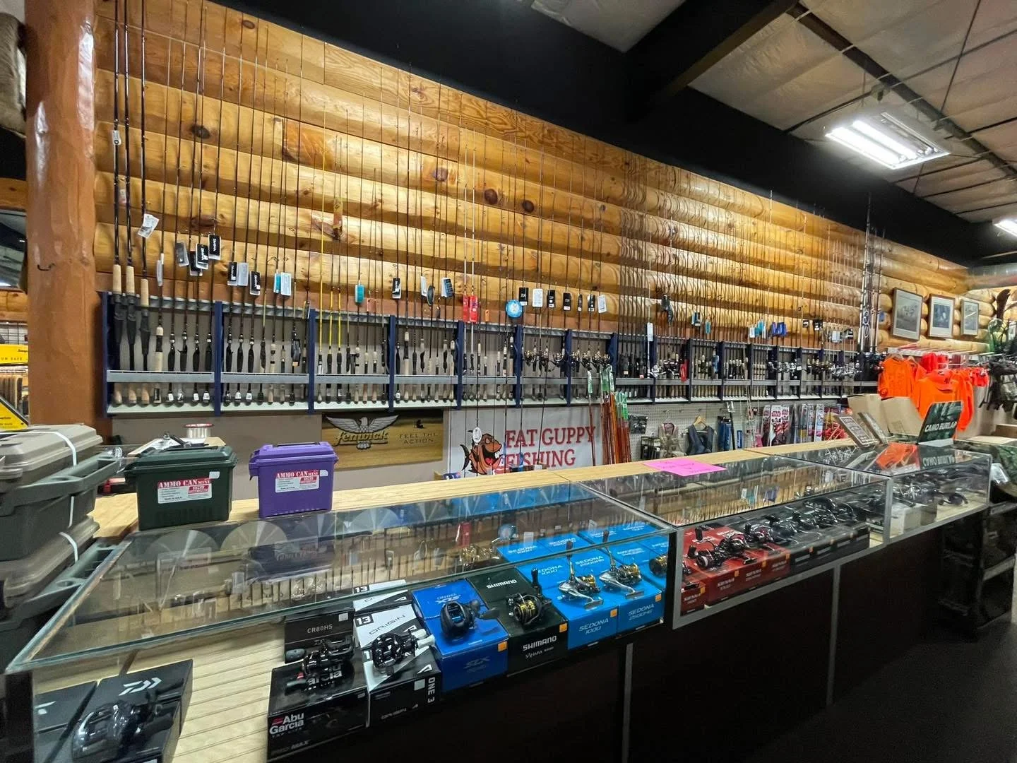 Fishing gear shop interior with fishing rods on the wall, tackle boxes and reels displayed on glass counter, and outdoor apparel hanging on the side.