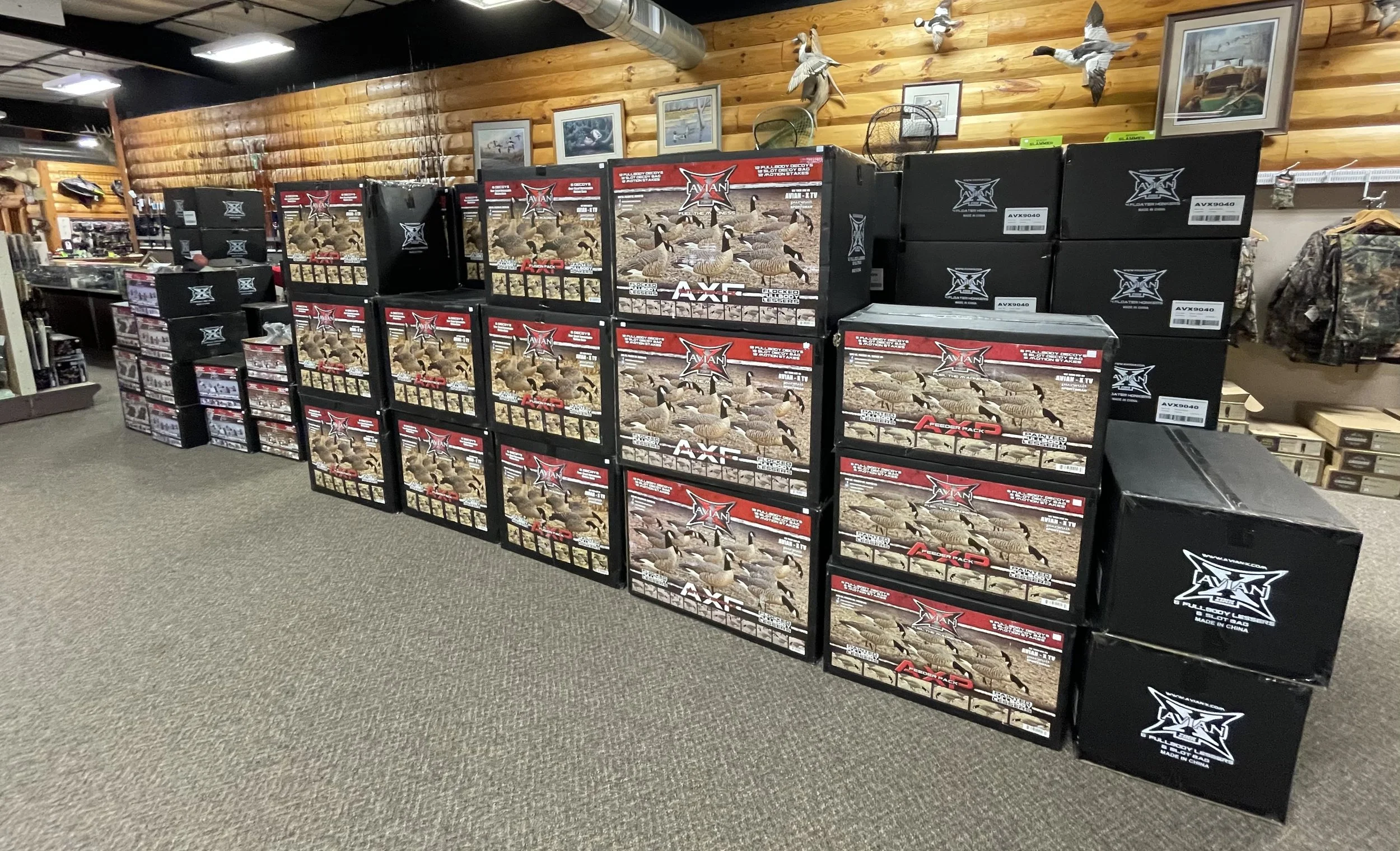 Stacked boxes of hunting decoys featuring ducks on the packaging, displayed on a carpeted floor inside a store with wood-paneled walls, framed pictures, and mounted ducks.