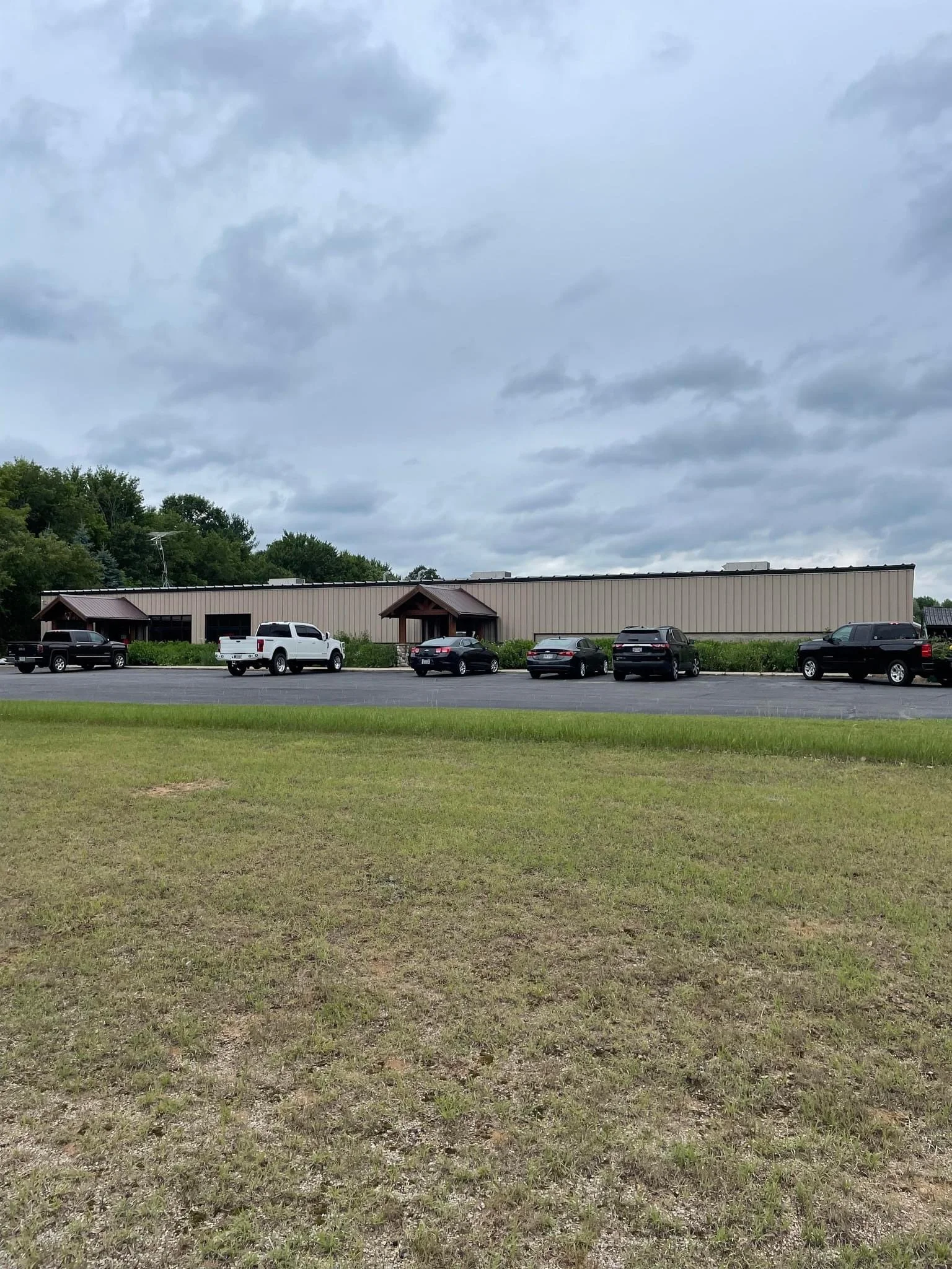 Parking lot with several cars parked in front of a large, beige industrial building under cloudy sky. Green grass in foreground.